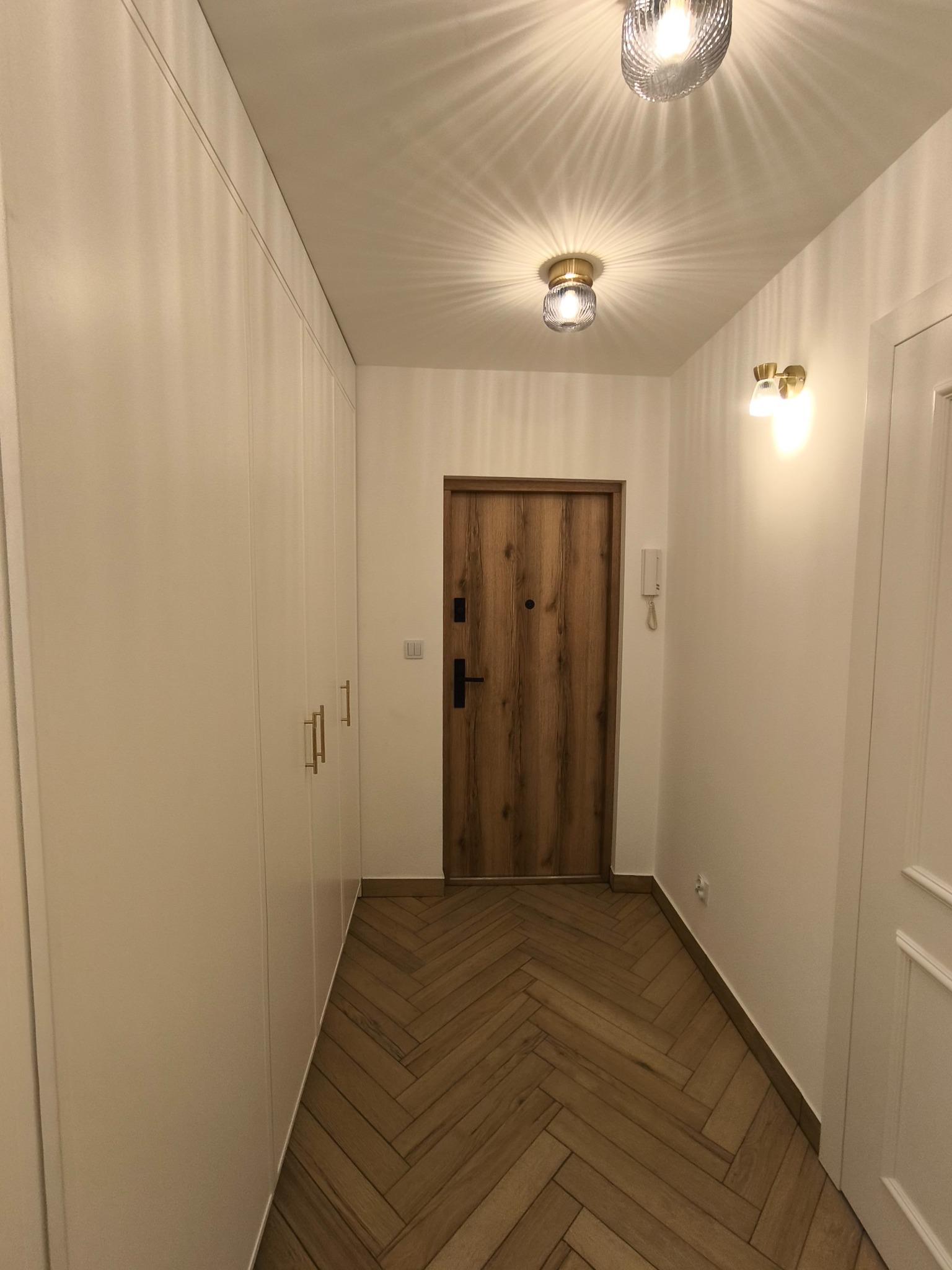 Przedpokój z parkietem ułożonym w jodełkę, biała zabudowa szafy z złotymi uchwytami, drewniane drzwi wejściowe z czarną klamką, oświetlenie sufitowe z dekoracyjnymi kloszami.