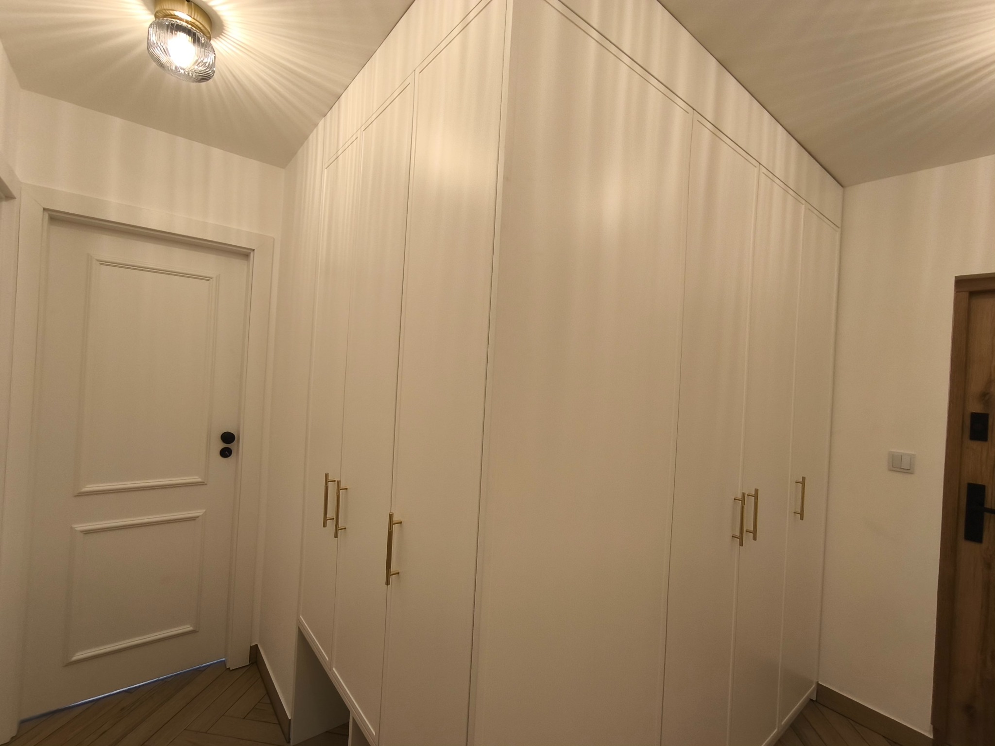 Wbudowana szafa z białymi frontami i złotymi uchwytami, dopasowana do skosu sufitu, widoczna z perspektywy podłogi w wykończonym wnętrzu.