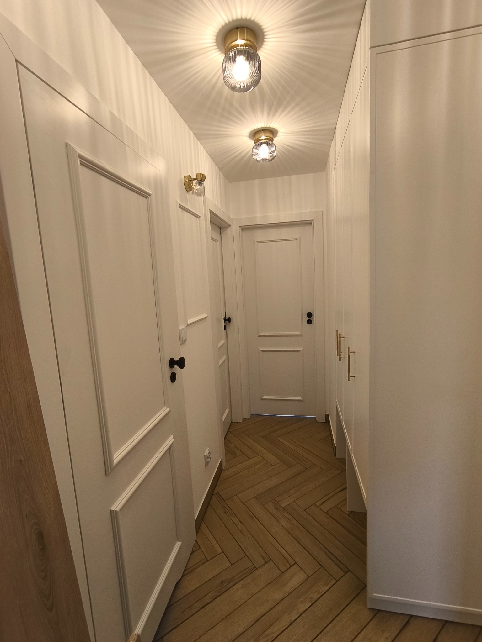 Korytarz w jasnych barwach z parkietem ułożonym w jodełkę, białymi drzwiami z czarnymi klamkami i złotymi lampami sufitowymi z dekoracyjnymi kloszami.