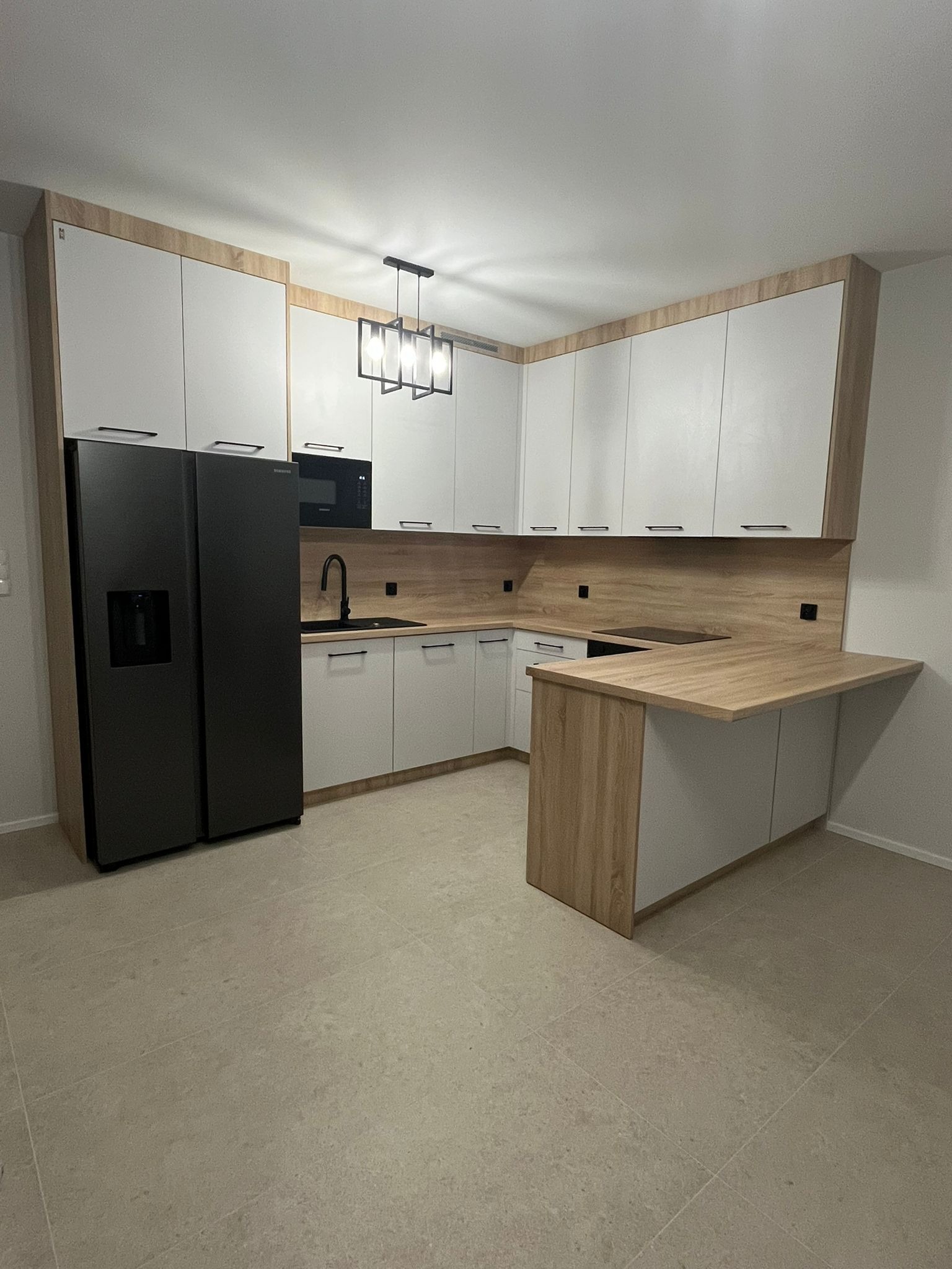 Nowoczesna kuchnia z białymi szafkami, drewnianym blatem i czarnym sprzętem AGD, oświetlona geometryczną lampą wiszącą.