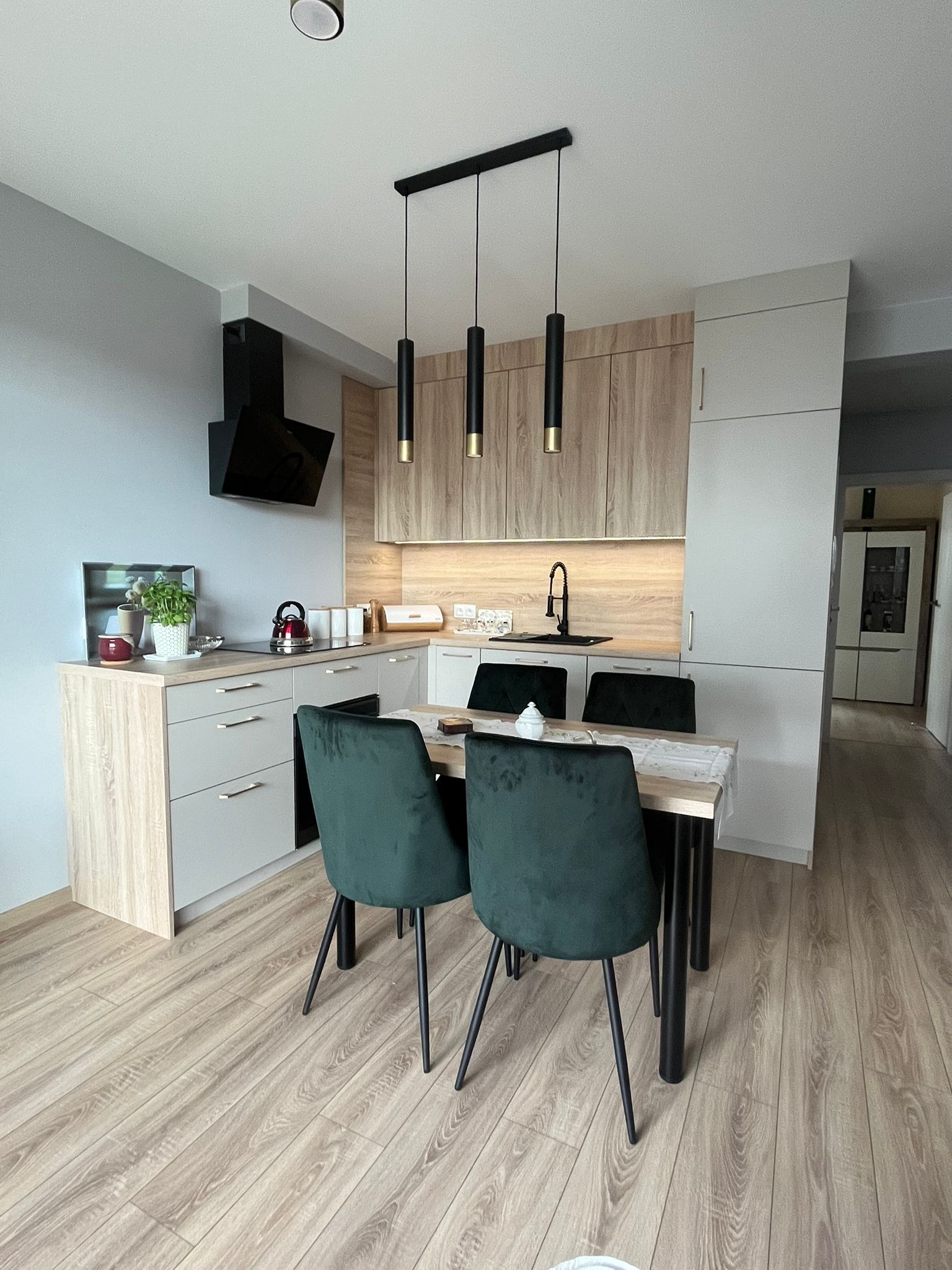 Nowoczesna aranżacja kuchni z jadalnią w Gdyni: jasne meble z drewnianymi akcentami, czarne lampy wiszące nad stołem z zielonymi krzesłami, drewniana podłoga.