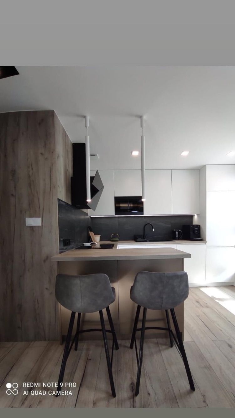 Nowoczesna kuchnia z wyspą, białe szafki, czarny okap i backsplash, drewnopodobna okładzina ściany, dwa szare hokery z czarnymi nogami, minimalistyczne oświetlenie.