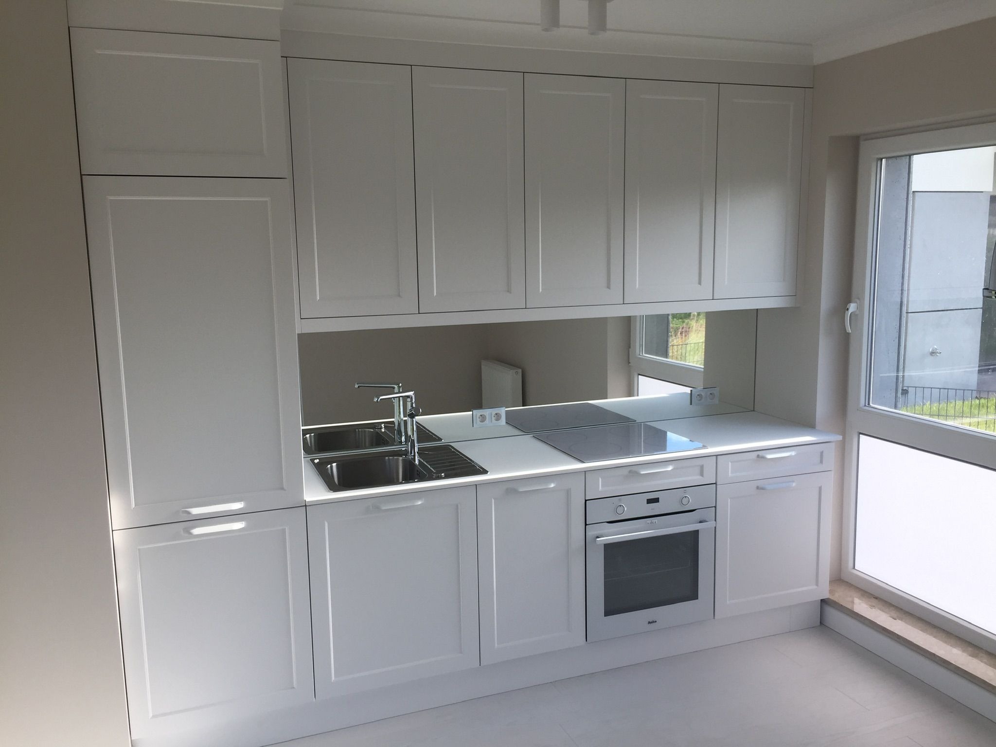 Nowoczesna, minimalistyczna kuchnia w bieli z lustrzanym panelem nad blatem, widoczne dwukomorowy zlew, płyta indukcyjna i piekarnik Amica, oświetlenie LED pod szafkami.