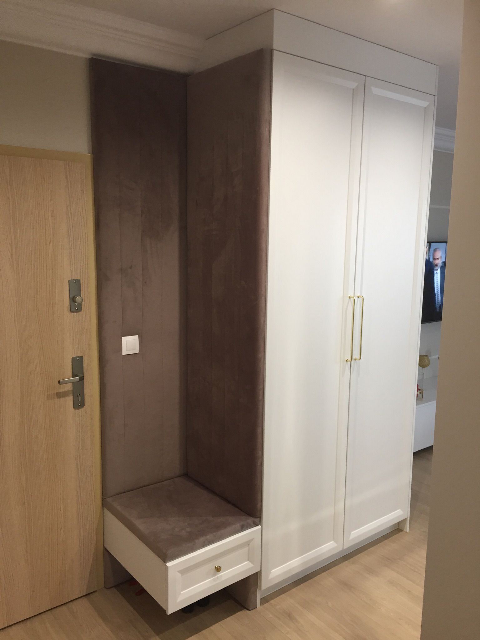 Wnęka z tapicerowanym siedziskiem i panelem obok drzwi wejściowych, zintegrowana z białą szafą w przedpokoju, drewniana podłoga.