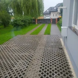 Ułożona nawierzchnia z ażurowych płyt betonowych na podjeździe do domu, z widocznymi pasami trawy między płytami, prowadząca do bramy wjazdowej i ogrodzenia z ceglanymi słupkami.