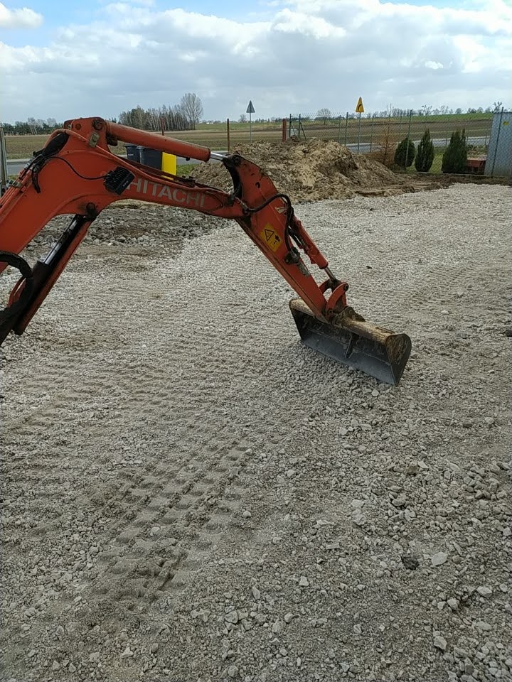 Pomarańczowa mini koparka Hitachi na placu pokrytym drobnym kamieniem, z hałdą ziemi w tle i znakami drogowymi.