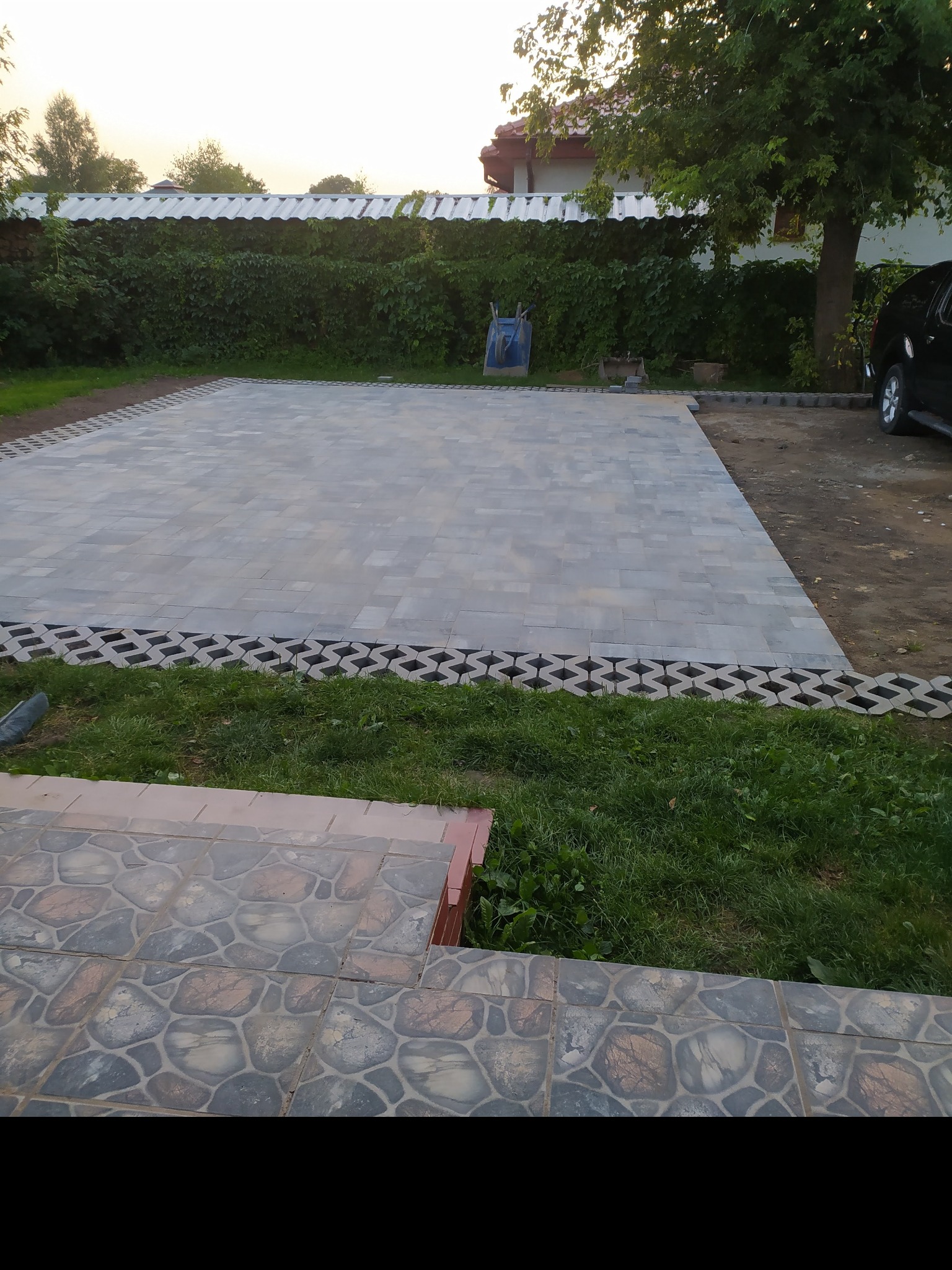 Ukończona powierzchnia utwardzona z szarej kostki brukowej na posesji, z obrzeżem z ażurowych płyt, w tle zielony żywopłot i drzewo.
