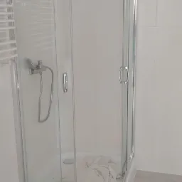 Narożna kabina prysznicowa z chromowanymi profilami i szklanymi drzwiami przesuwnymi, zamontowana w jasnej łazience z szarymi płytkami podłogowymi i grzejnikiem drabinkowym.