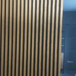 Drewniane lamele na czarnym tle tworzące ściankę działową, widoczne oświetlenie sufitowe i fragment łazienki w tle.