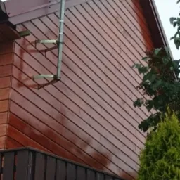 Fragment ściany budynku obłożonej brązowymi panelami drewnianymi z zamontowaną drabiną do anteny satelitarnej, widoczne elementy dachu i fragment ogrodzenia.