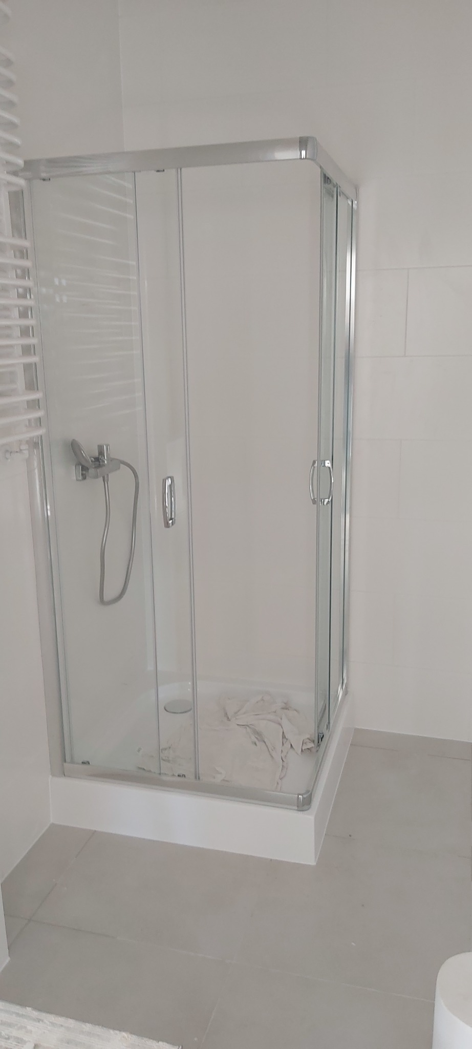 Narożna kabina prysznicowa z chromowanymi profilami i szklanymi drzwiami przesuwnymi, zamontowana w jasnej łazience z szarymi płytkami podłogowymi i grzejnikiem drabinkowym.