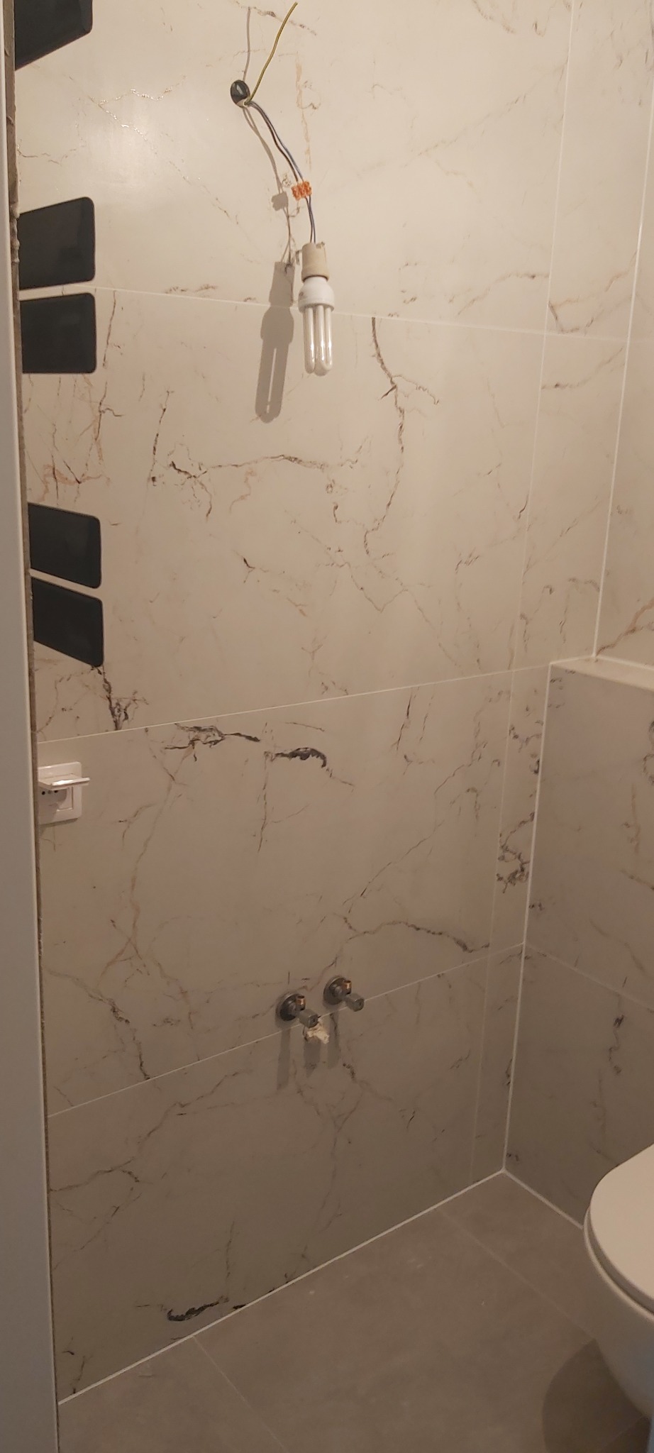 Ściana łazienki wyłożona płytkami imitującymi marmur, z widocznymi wystającymi przewodami elektrycznymi i żarówką, oraz fragmentem toalety; widoczne niedociągnięcia w wykończeniu.
