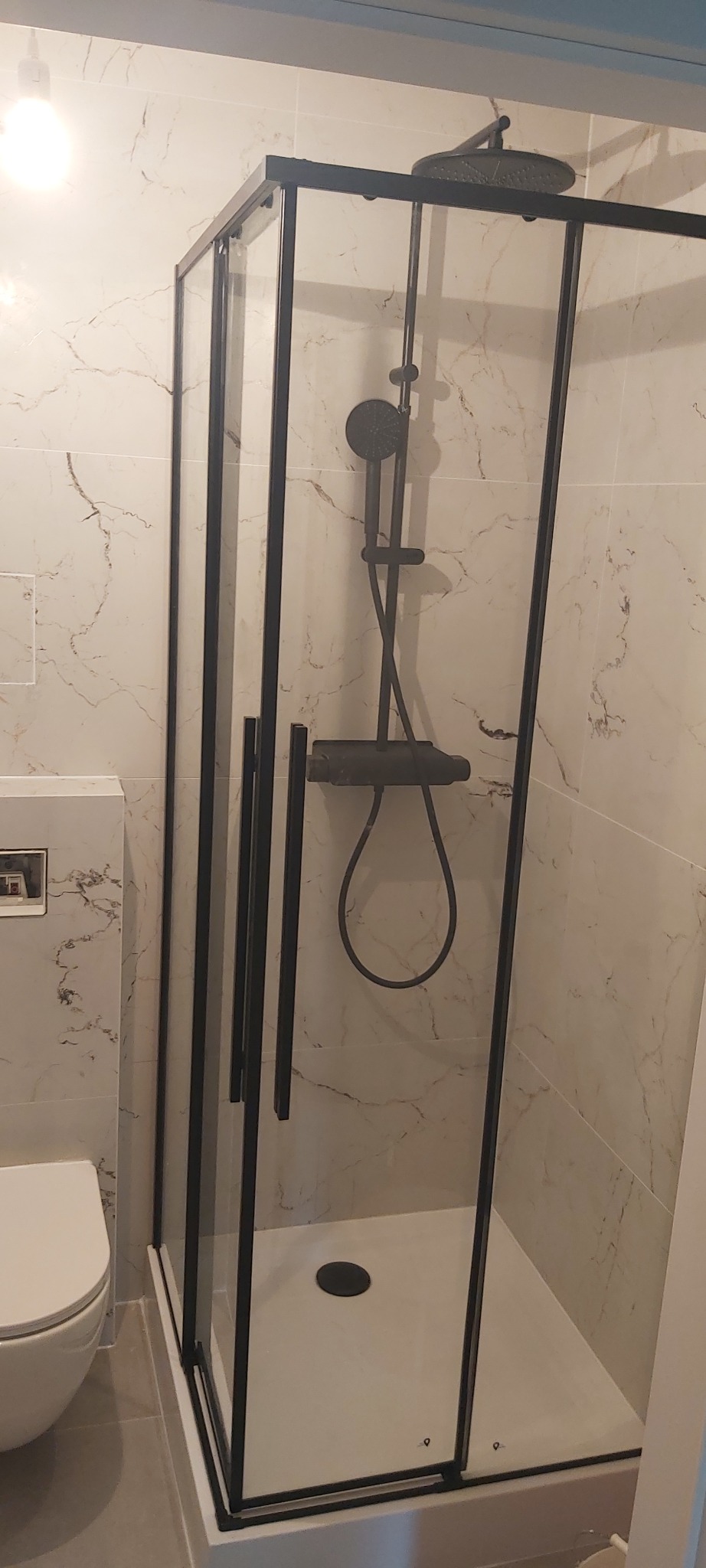 Nowoczesna kabina prysznicowa z czarną armaturą i ramą, biały brodzik, marmurowe płytki na ścianach, widoczna toaleta w tle.