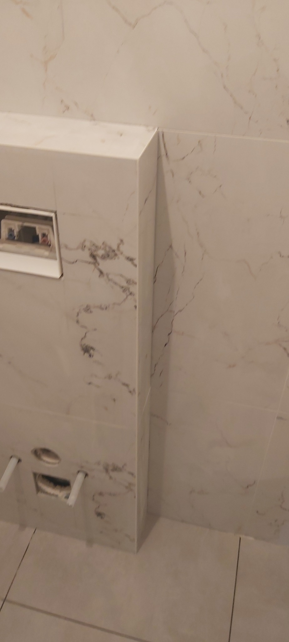 Ściana i podłoga wyłożone jasnymi płytkami imitującymi marmur, widoczna instalacja podtynkowego WC w trakcie montażu.