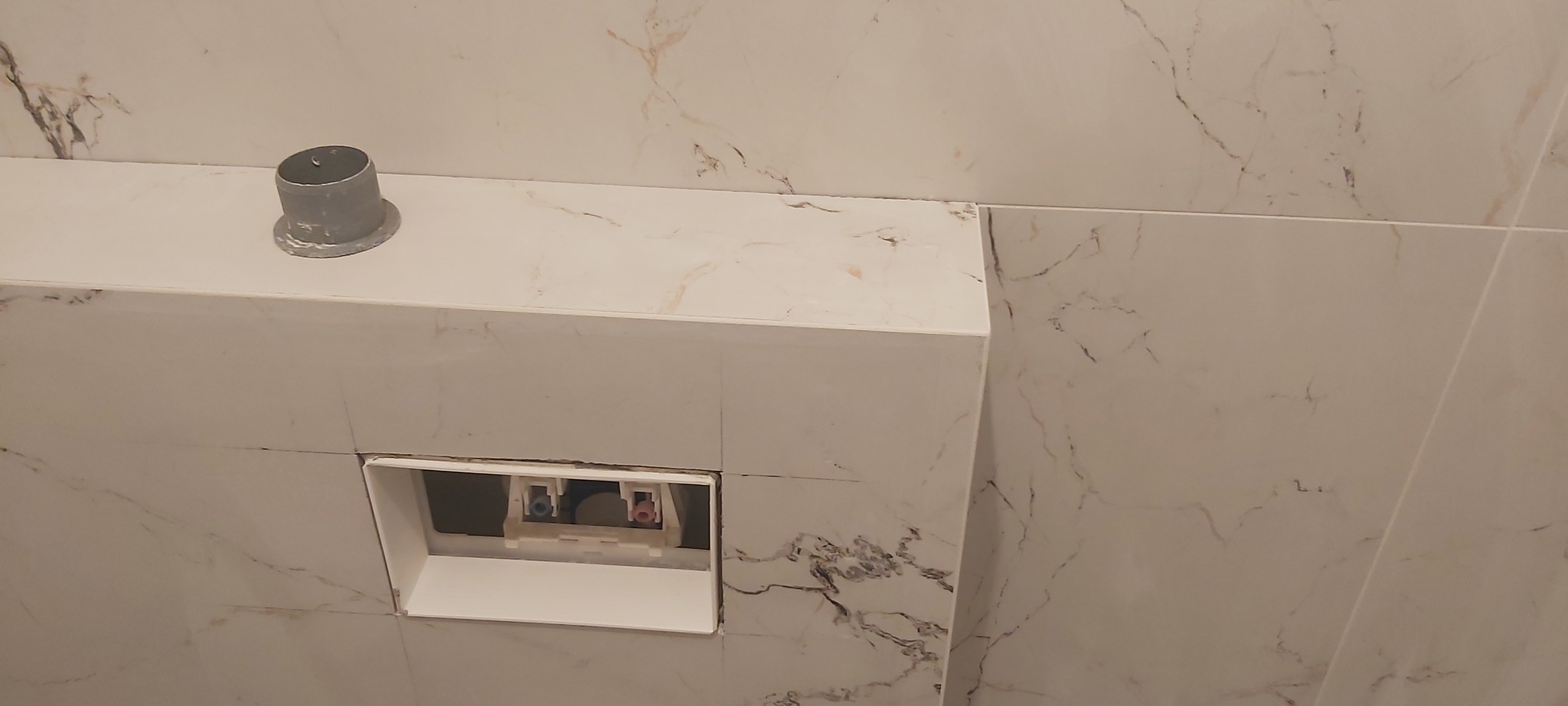 Kafelki imitujące marmur na zabudowie stelaża podtynkowego WC z widocznym otworem na przyciski spłukujące i wystającym elementem instalacji.
