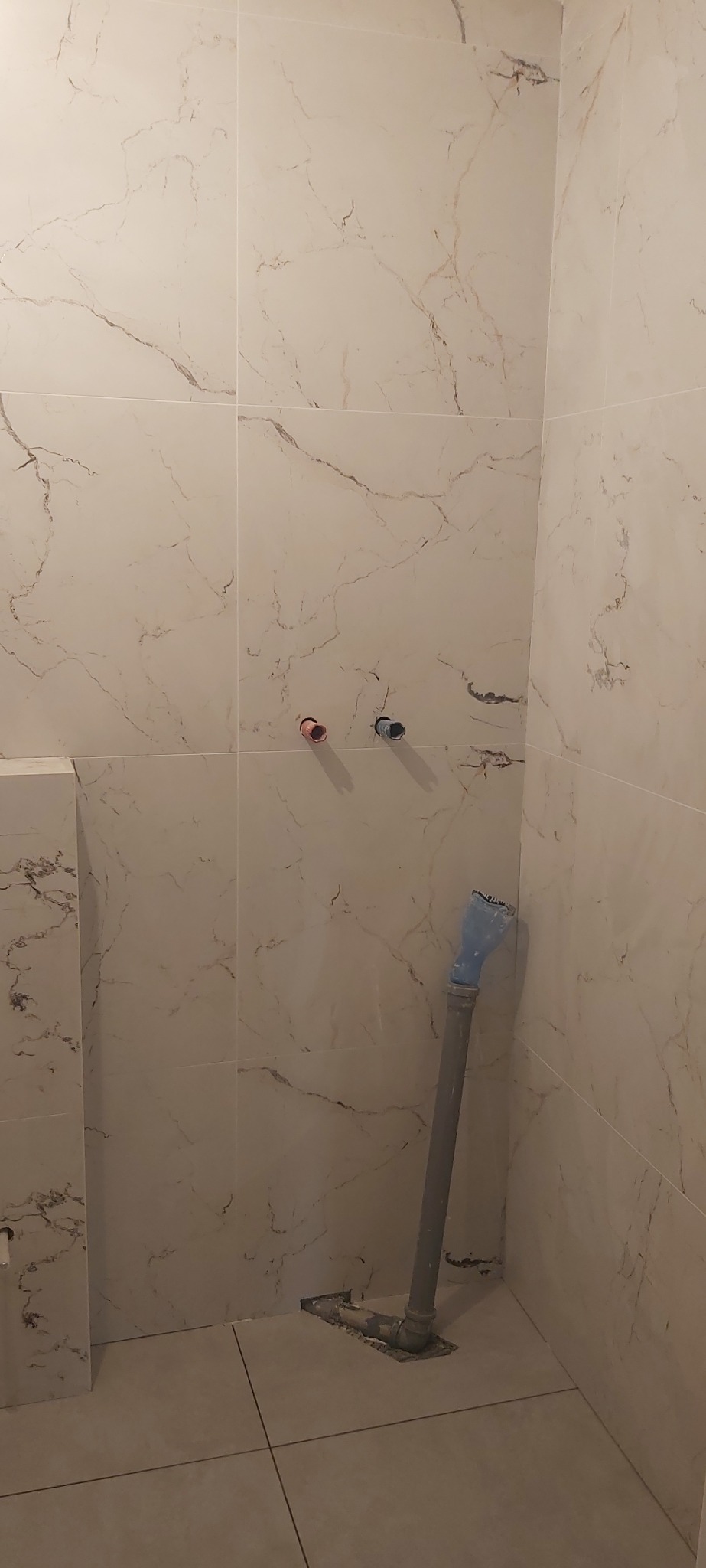 Ściana łazienki wyłożona jasnymi płytkami imitującymi marmur, widoczne wystające rury z ciepłą i zimną wodą oraz pion kanalizacyjny z niebieską końcówką, przy podłodze widoczne pozostałości po...