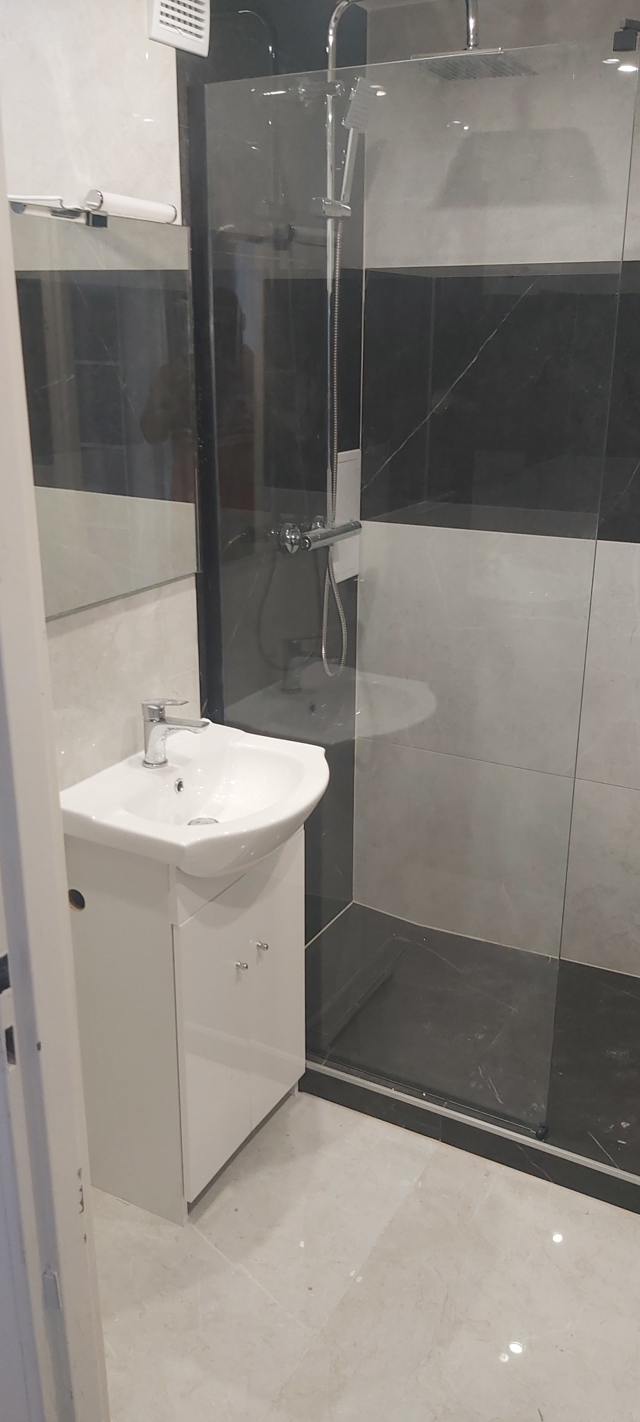 Nowoczesna łazienka z białą umywalką z szafką, kabiną prysznicową ze szklaną ścianą i czarno-białymi płytkami na ścianach oraz jasnymi płytkami na podłodze.