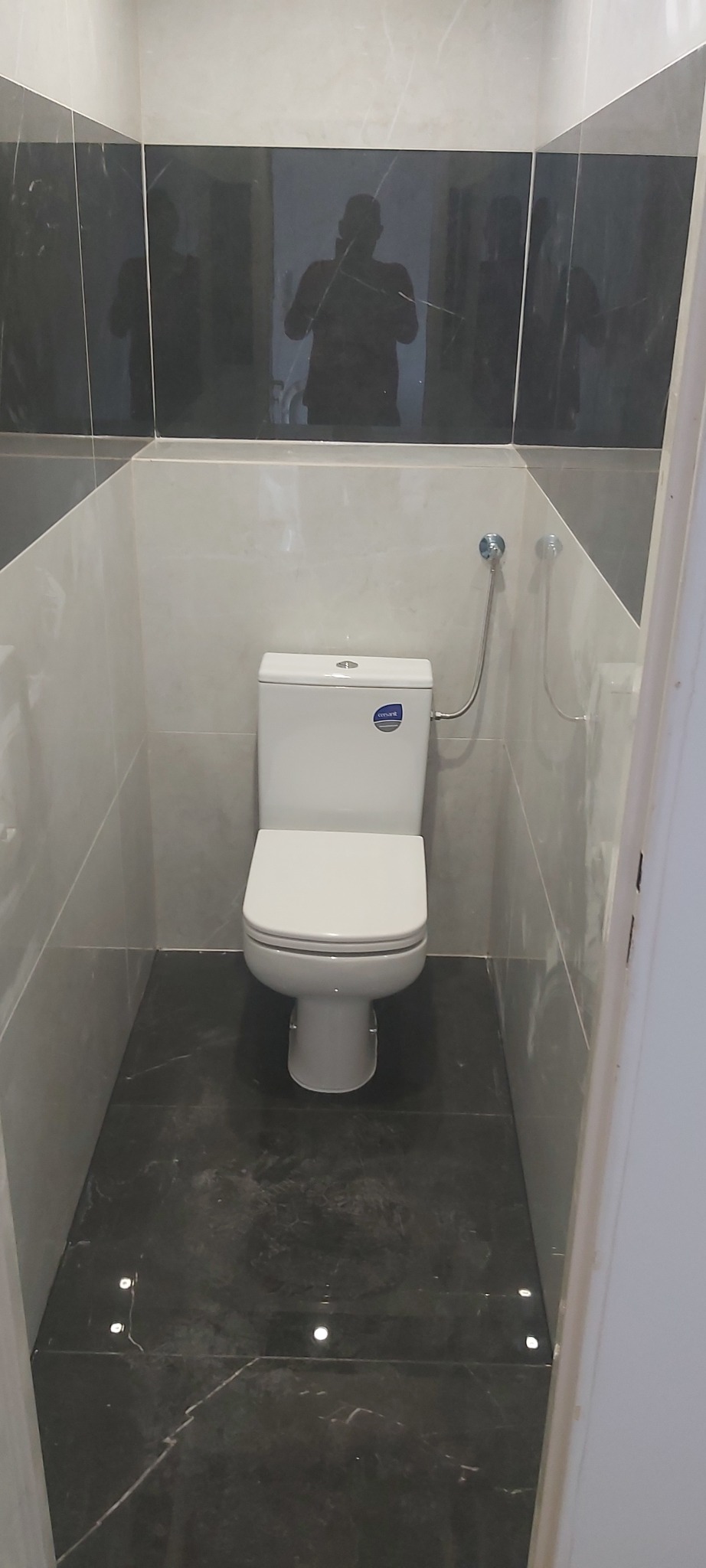 Nowoczesna toaleta z białym sedesem, szarymi i czarnymi płytkami na ścianach i podłodze, z widocznym odbiciem fotografa w czarnych płytkach.