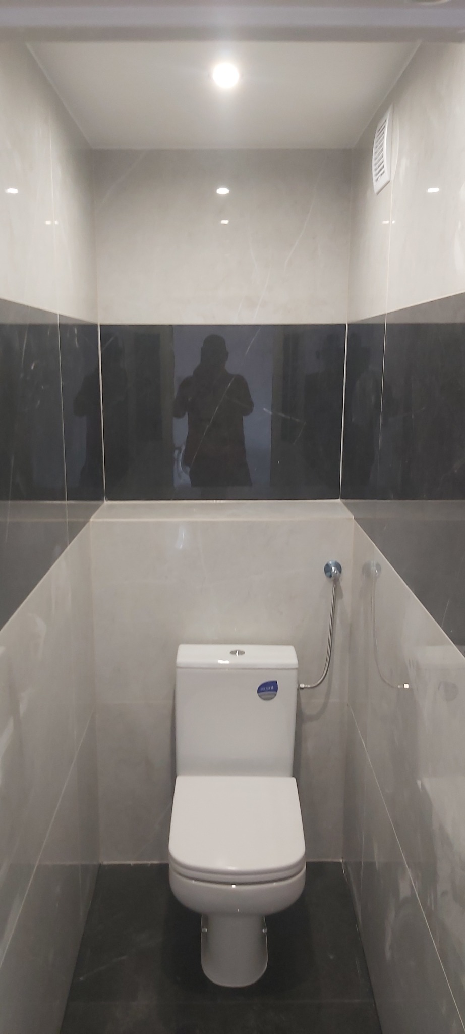 Wykończona toaleta z białym kompaktem WC, szarymi ścianami z marmurowym wzorem, czarnym pasem płytek na ścianie i odblaskiem fotografa w płytkach.