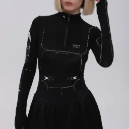 Modelka w czarnym topie z długim rękawem i czarnej spódnicy, prezentująca strój z kolekcji lookbook 2022 na jednolitym, jasnym tle.