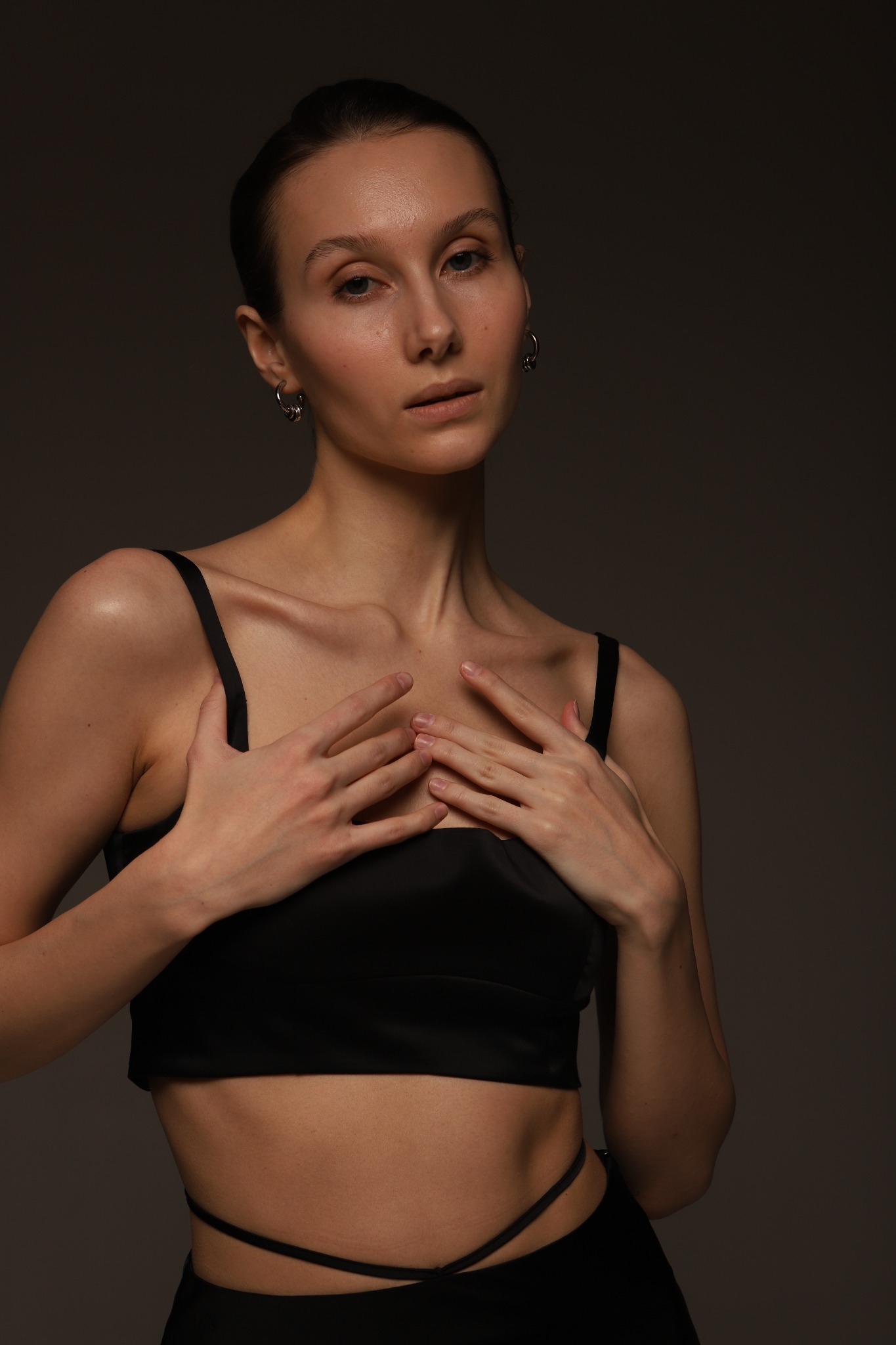 Portret kobiety w czarnym topie i spodniach, z rękami na dekolcie, subtelne kolczyki koła, ciemne tło, oświetlenie studyjne.