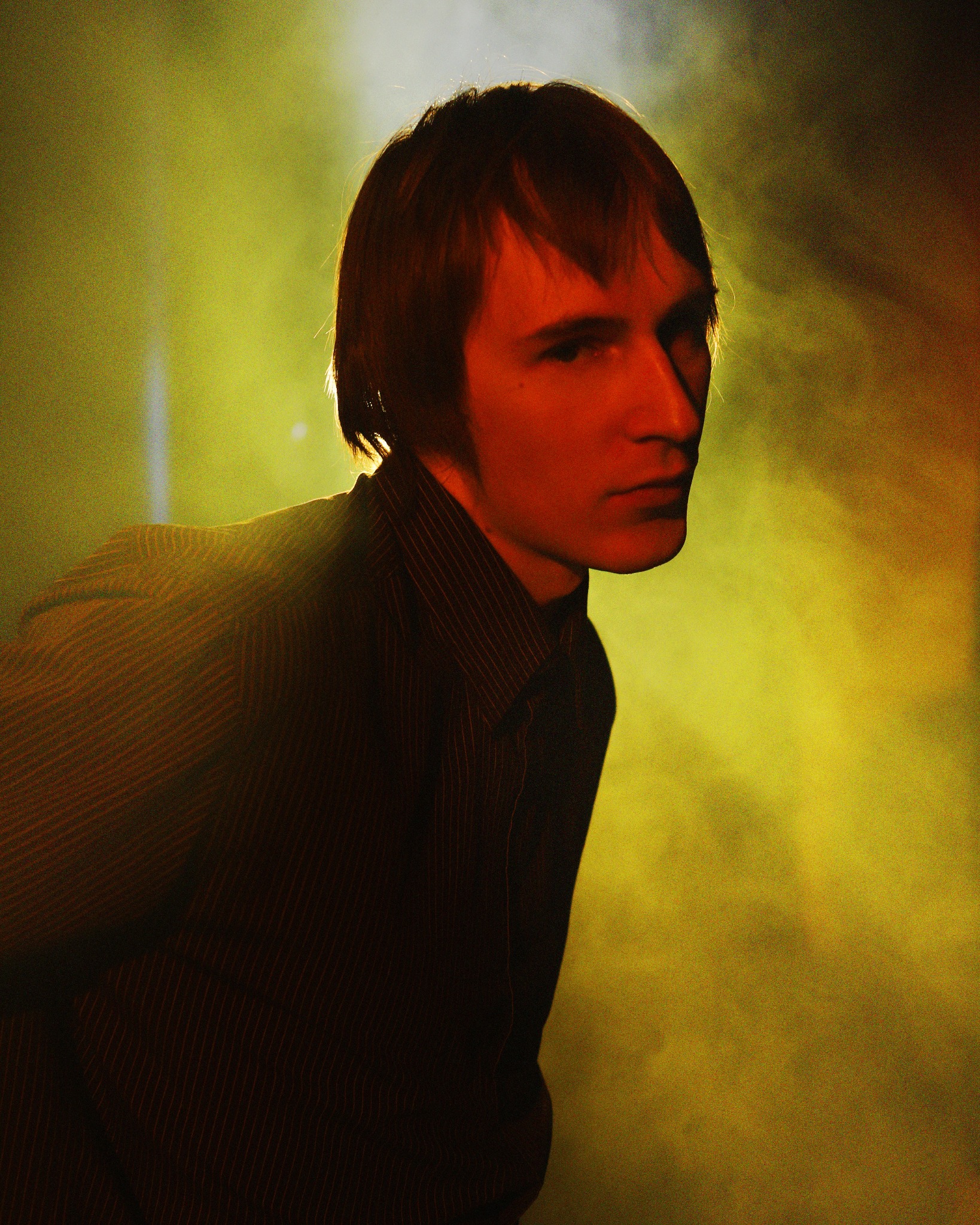 Portret młodego mężczyzny w prążkowanej koszuli, oświetlonego żółto-pomarańczowym światłem w zadymionym otoczeniu, z profilu.