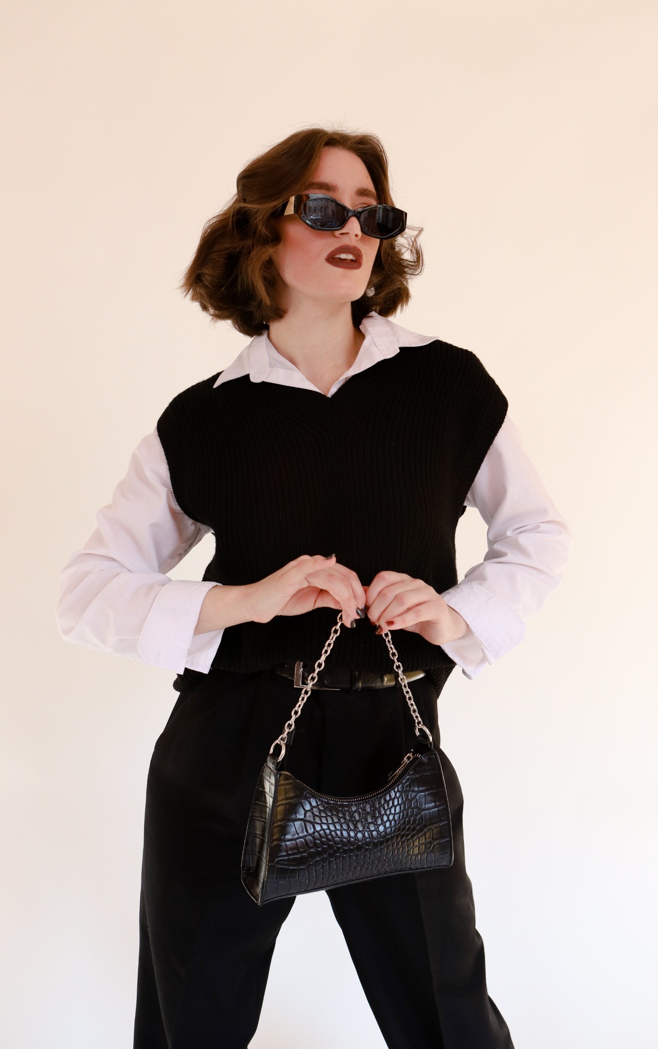 Kobieta w stylizacji retro: czarny sweter bez rękawów, biała koszula, czarne spodnie i torebka ze wzorem skóry krokodyla, pozuje w okularach przeciwsłonecznych.