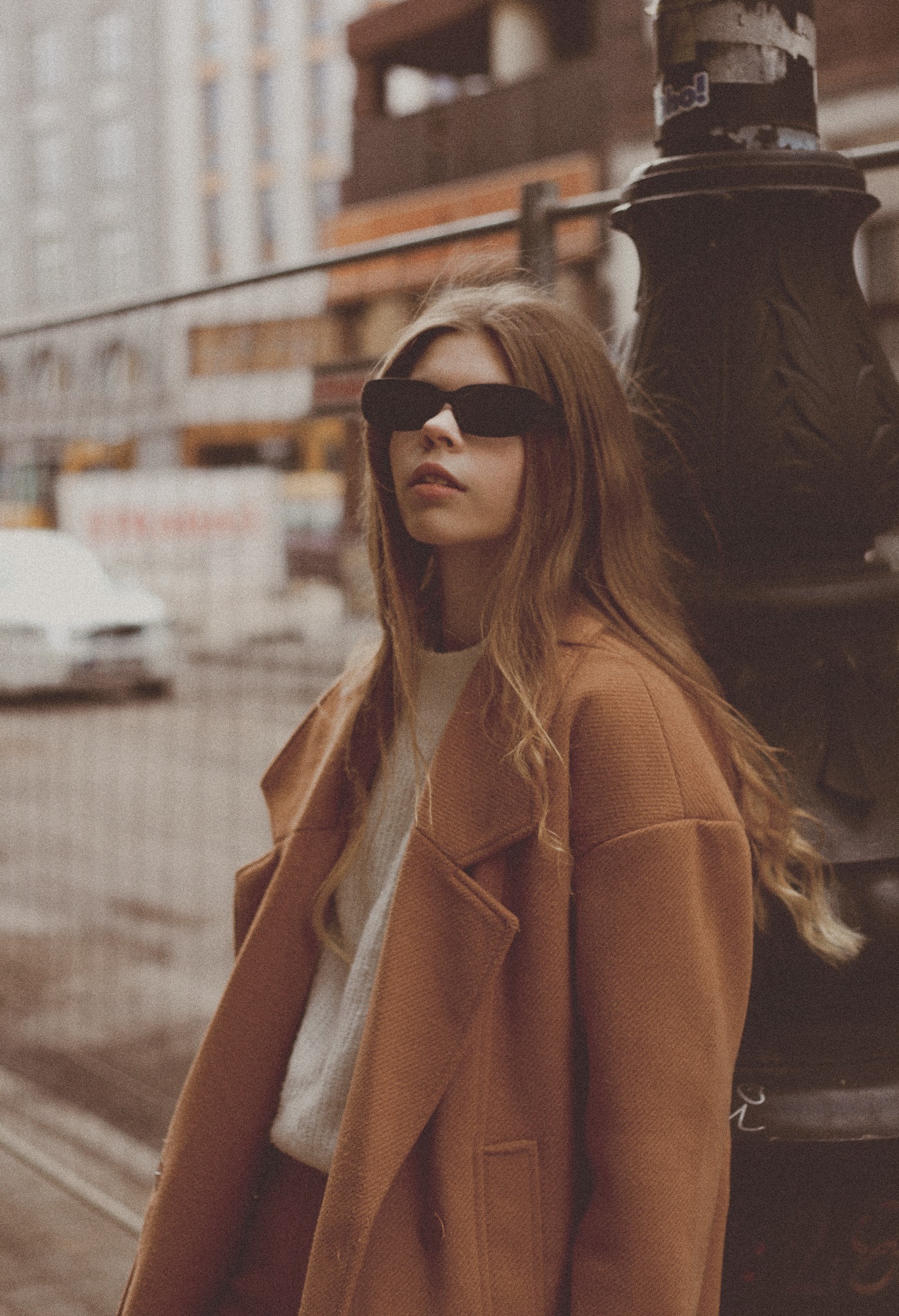 Portret młodej kobiety w brązowym płaszczu i okularach przeciwsłonecznych, opierającej się o latarnię w miejskim krajobrazie Wrocławia. W tle rozmyte budynki i przejeżdżające samochody.