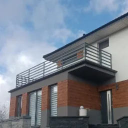 Nowoczesny dom z balkonem zabezpieczonym poziomą, szarą balustradą. Elewacja z elementami imitującymi drewno. Schody wejściowe wyłożone ciemną kostką.