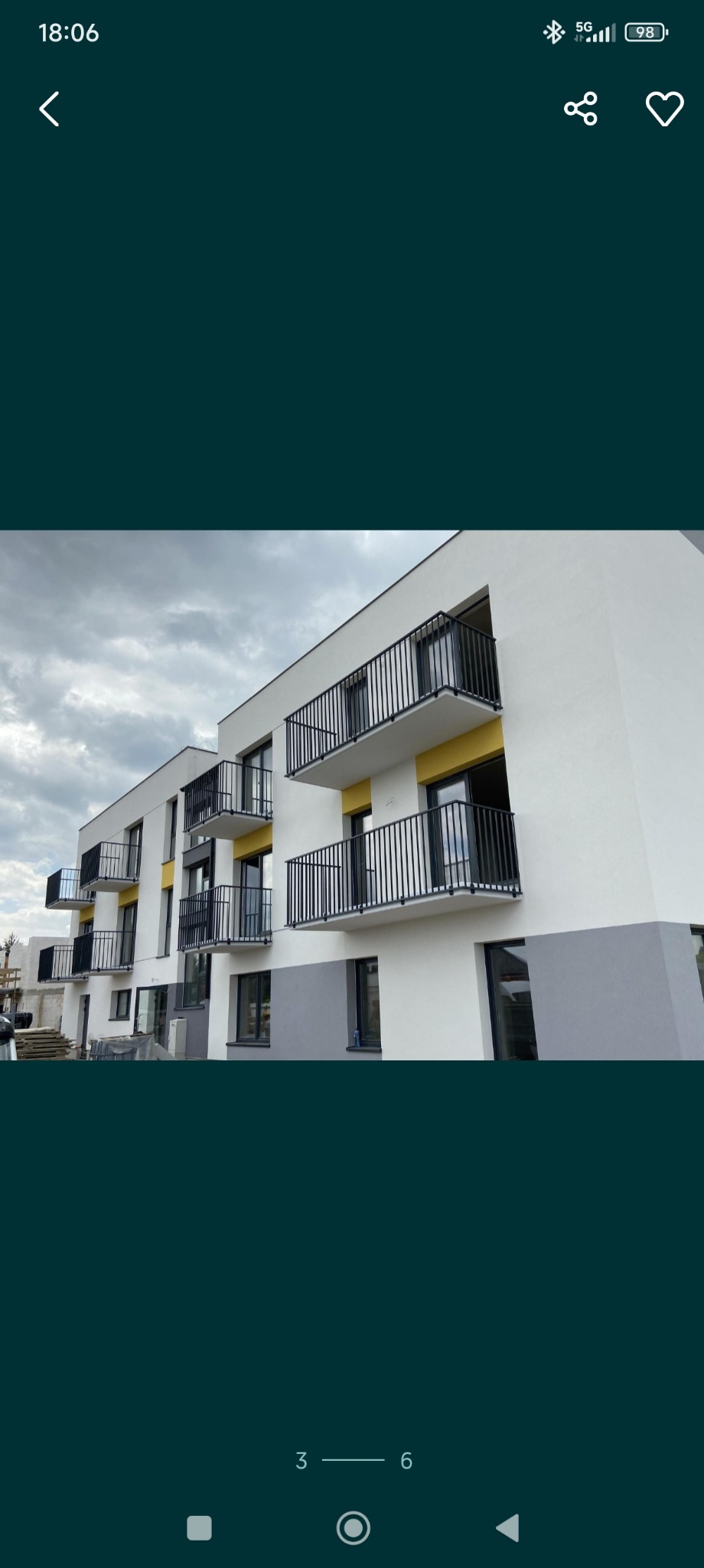 Nowoczesny budynek z balkonami z czarnymi barierkami. Żółte akcenty na elewacji i szare cokoły. Częściowo widoczne materiały budowlane.