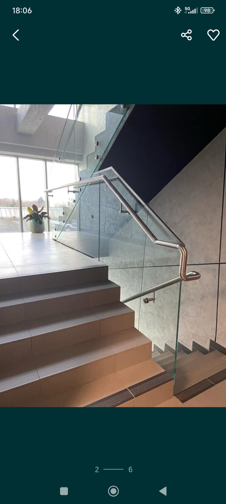 Nowoczesne schody z jasnymi stopniami, szklaną balustradą i metalową poręczą. Widok z dołu schodów, w tle okno i roślina doniczkowa.