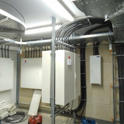 Elektro-Atom Tomasz Paciuch - Rozbudowana instalacja elektryczna w budynku: kable w korytach, białe skrzynki rozdzielcze, metalowe wsporniki. Widoczne elementy montażowe i narzędzia.