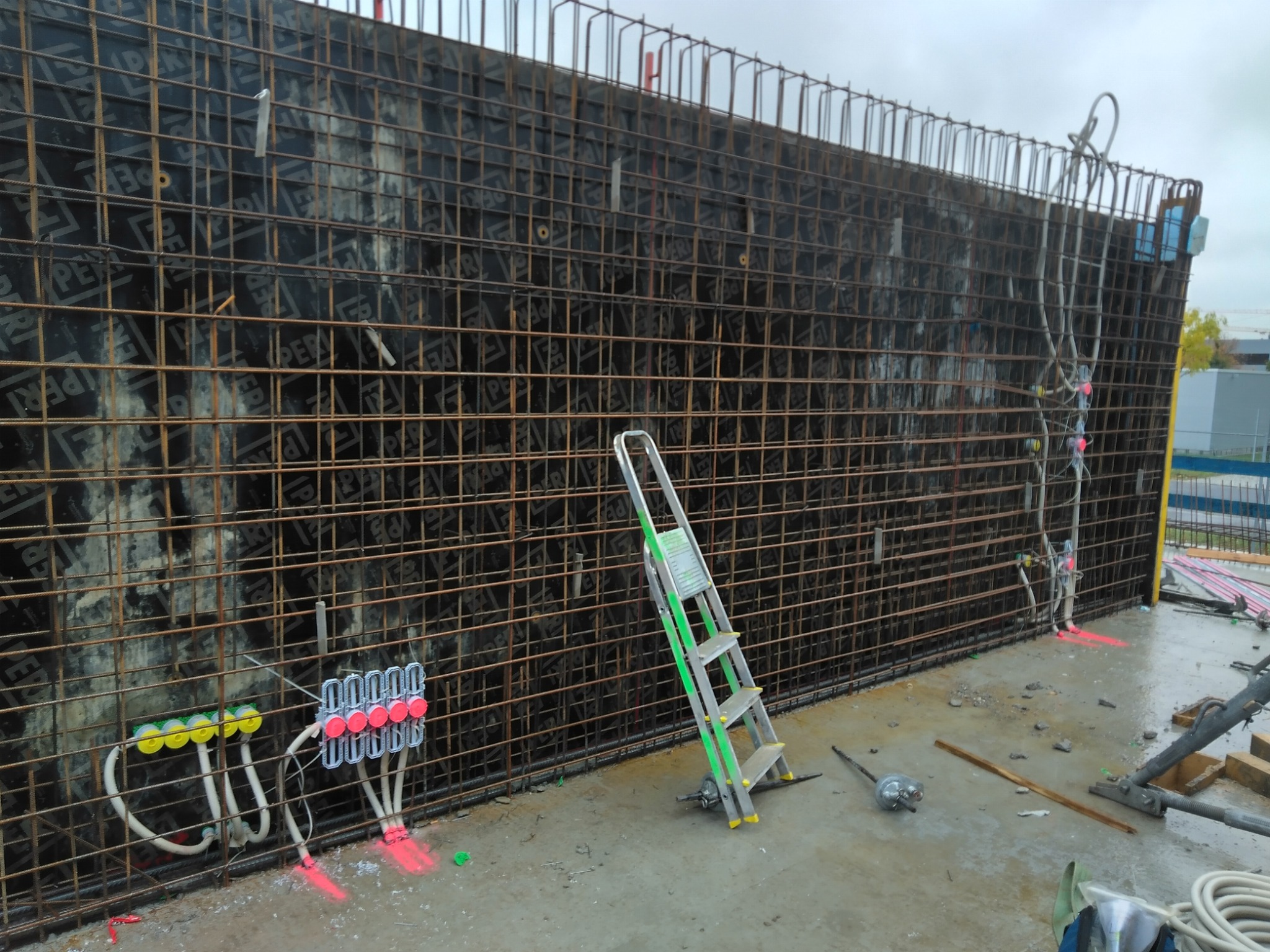 Zbrojenie ściany z widocznymi puszkami instalacyjnymi i przewodami. Na mokrym betonie stoi drabina. Widoczne elementy instalacji odgromowej.