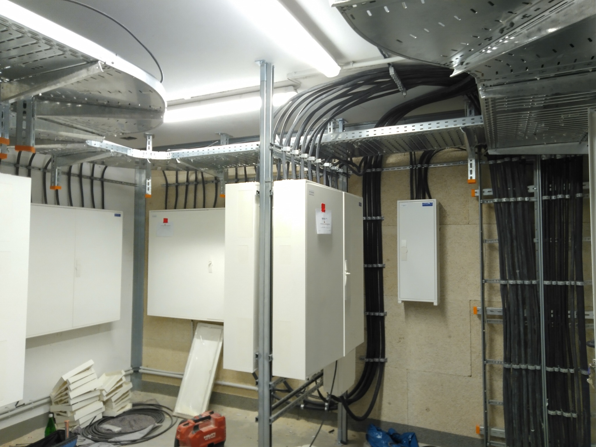 Rozbudowana instalacja elektryczna w budynku: kable w korytach, białe skrzynki rozdzielcze, metalowe wsporniki. Widoczne elementy montażowe i narzędzia.