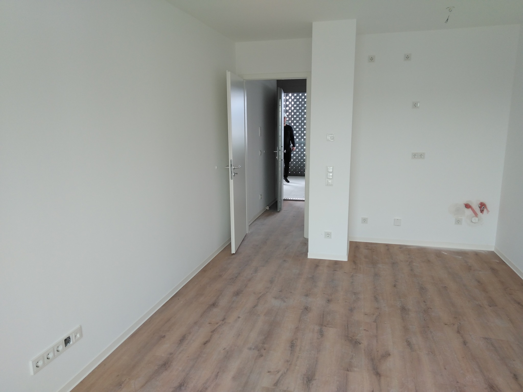 Nowe mieszkanie z drewnianą podłogą, otwartymi drzwiami i widocznymi gniazdkami elektrycznymi na ścianach. W tle widoczna osoba.