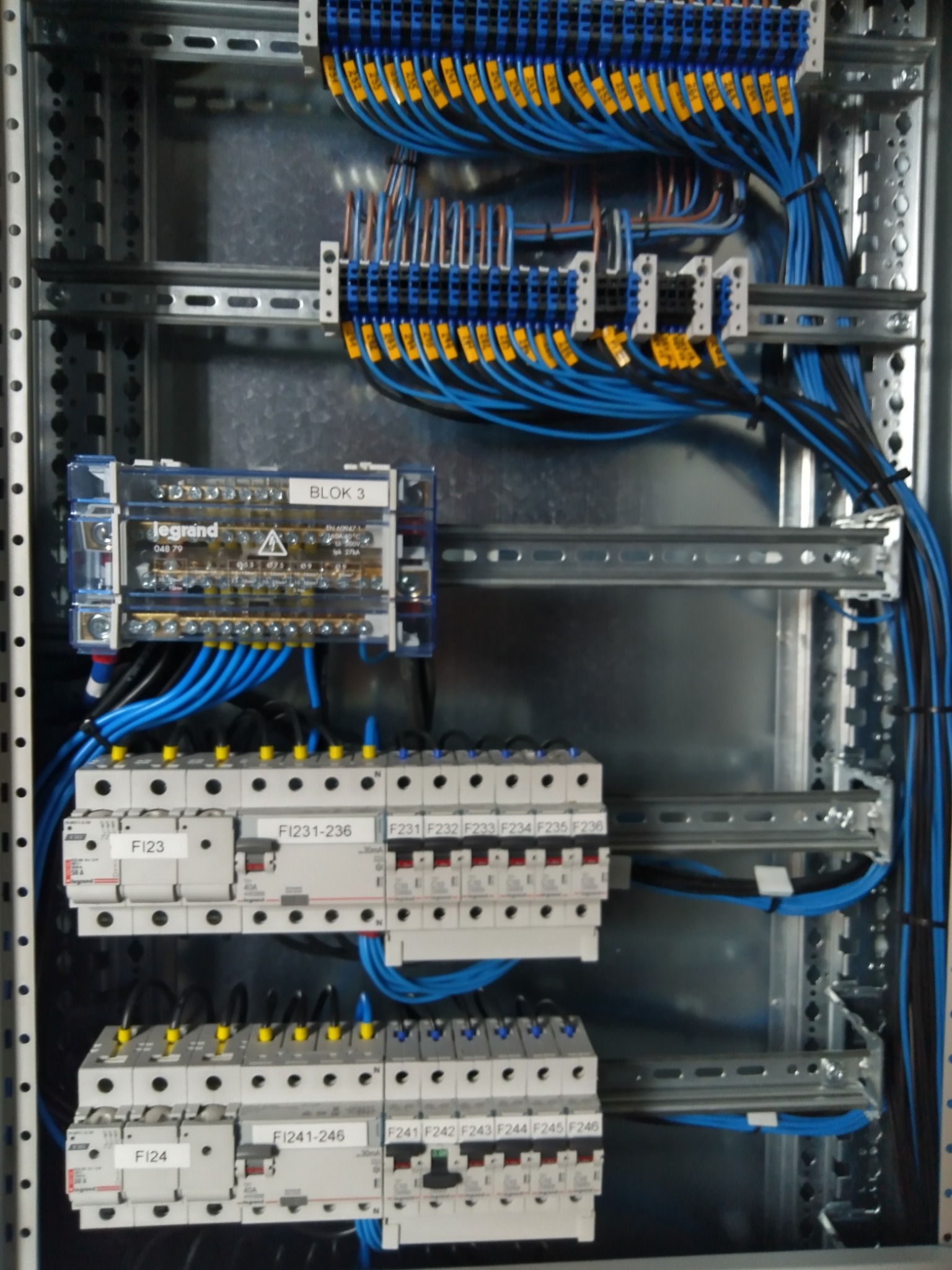 Rozdzielnia elektryczna z precyzyjnie ułożonymi niebieskimi przewodami, oznaczonymi żółtymi etykietami, zamontowana w metalowej obudowie z szynami DIN.