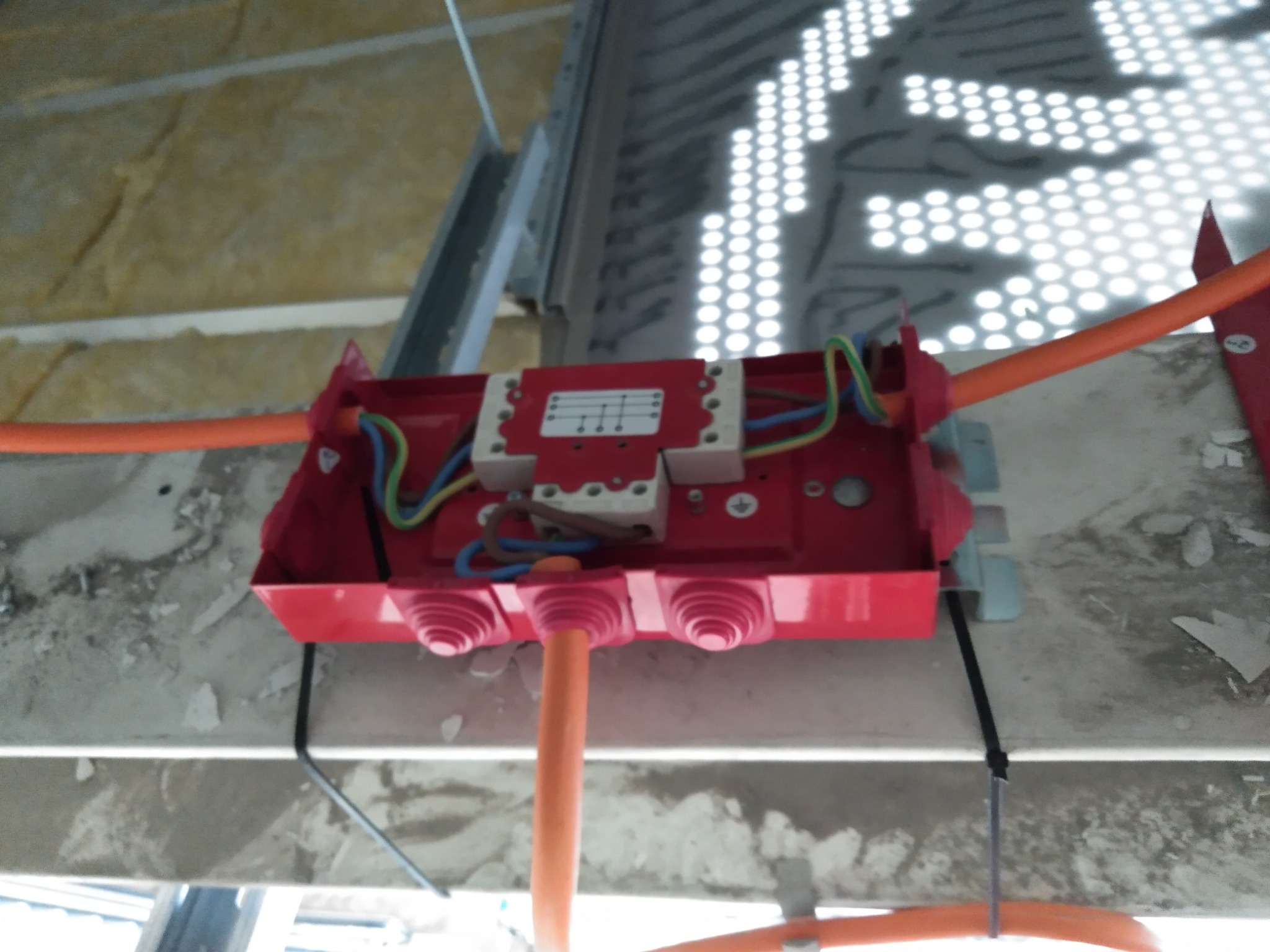 Czerwona puszka elektryczna z przewodami zamontowana na suficie podwieszanym z oświetleniem rastrowym. Widoczne kable w kolorach: pomarańczowy, niebieski, zielono-żółty.