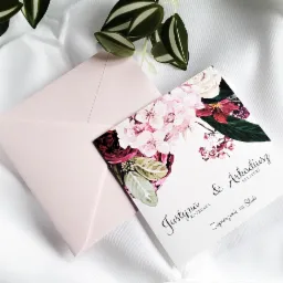 Eleganckie zaproszenie ślubne z motywem kwiatowym i różową kopertą, ułożone na białej, delikatnie pogniecionej tkaninie, z dodatkiem zielonej rośliny.