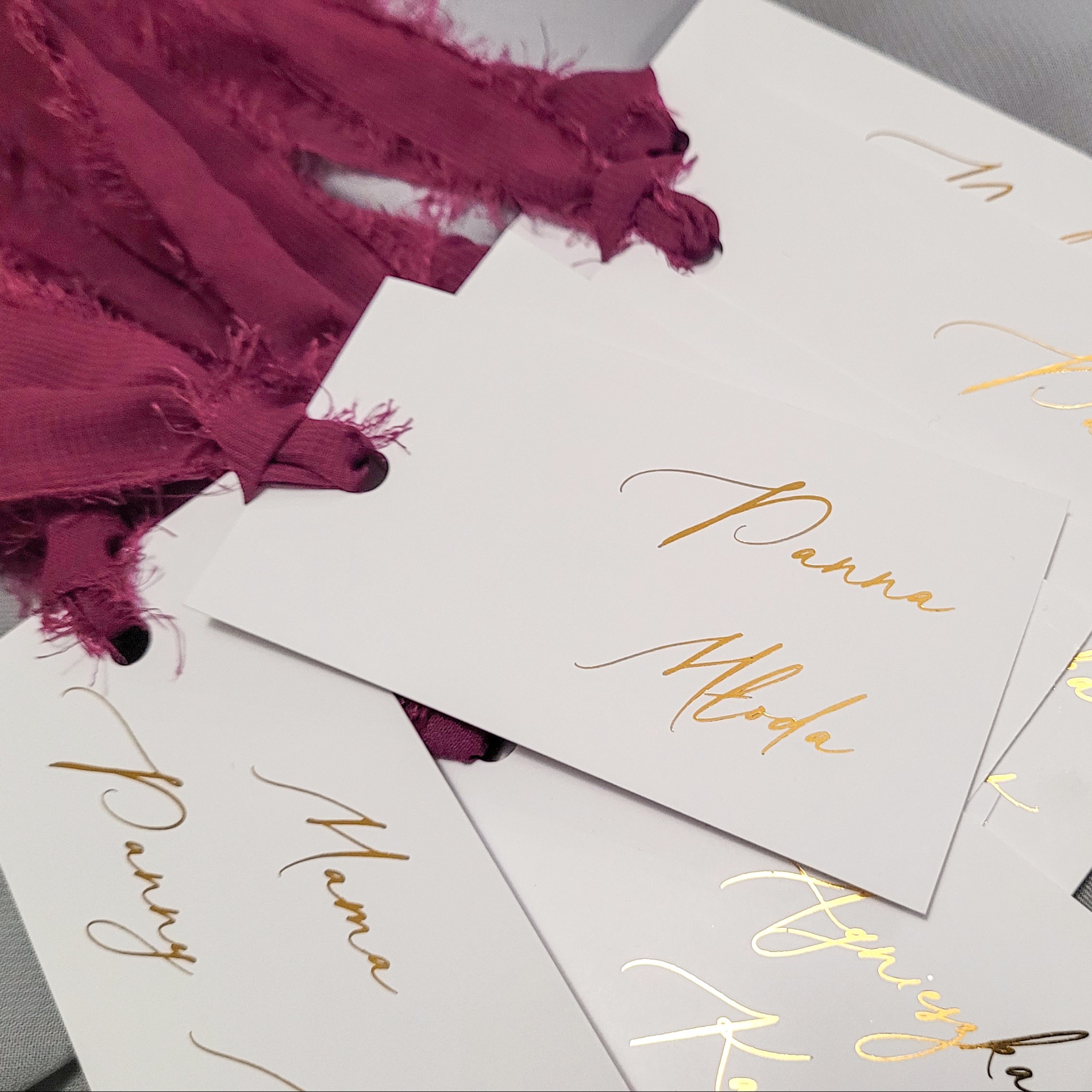 Stos białych, eleganckich winietek z imionami wypisanymi złotą kaligrafią, przewiązanych bordową, postrzępioną wstążką.