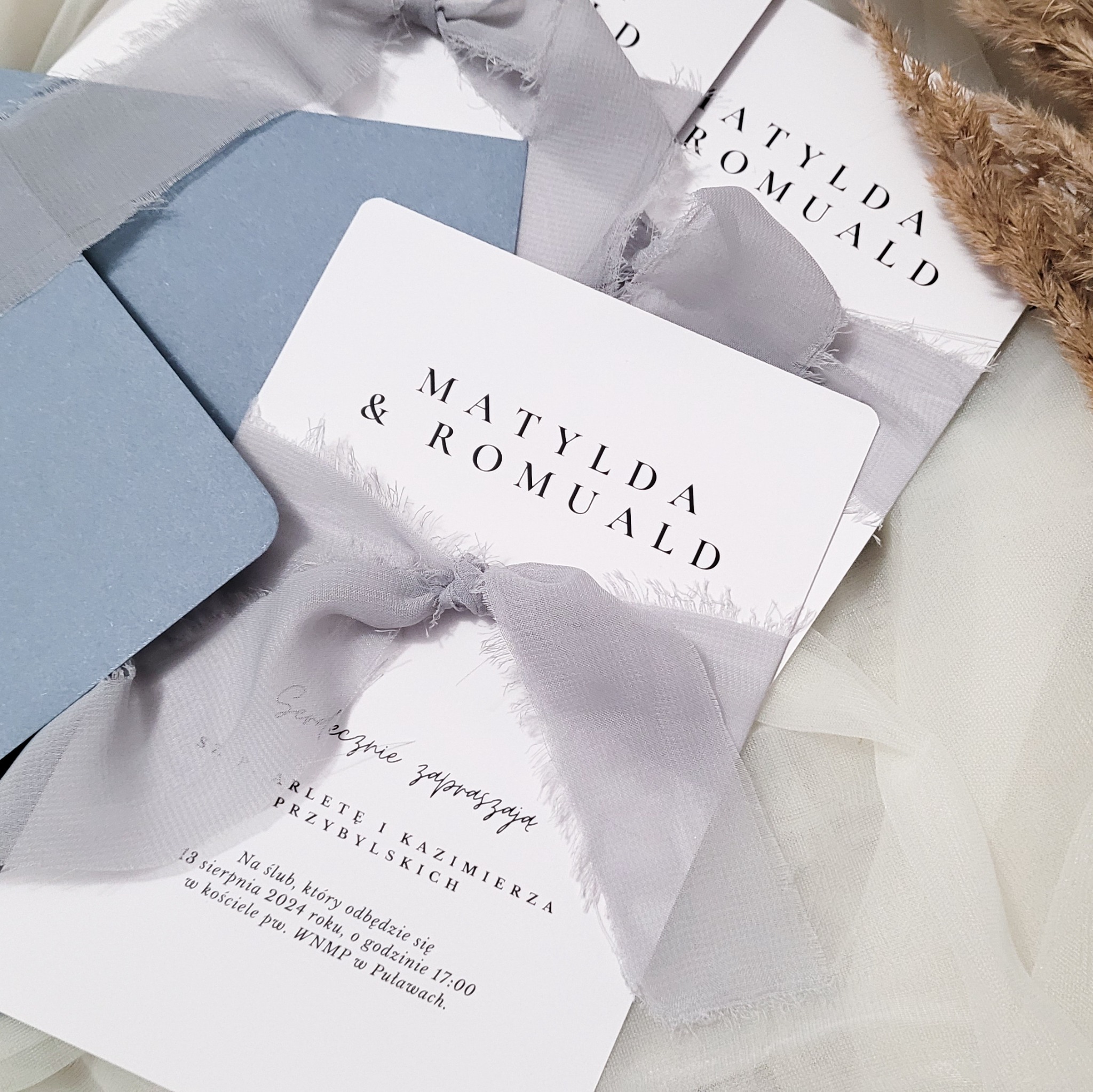 Eleganckie zaproszenia ślubne z imionami Matylda i Romuald, przewiązane delikatną szarą wstążką, obok niebieska koperta i suszone trawy na kremowym tle.