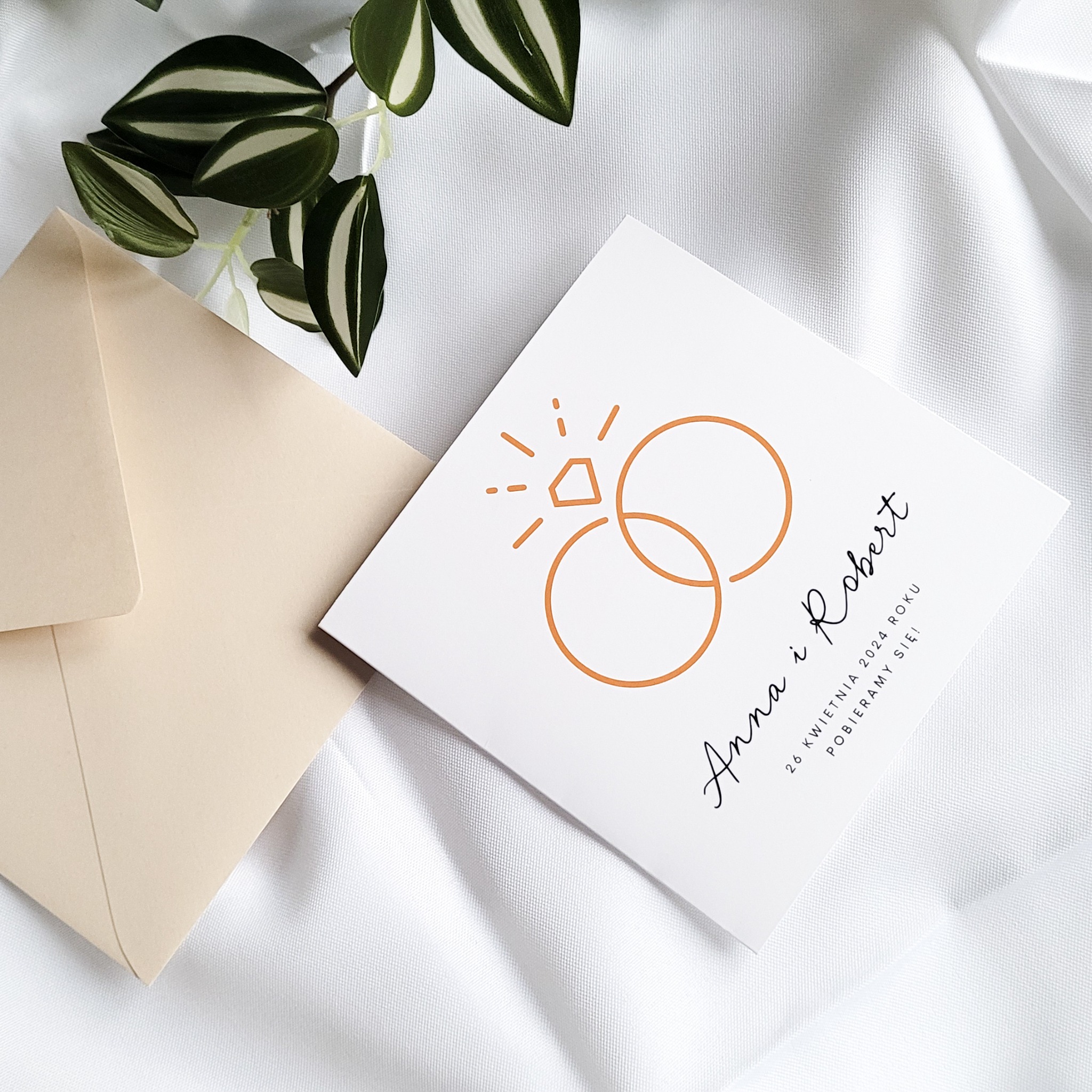 Eleganckie zaproszenie ślubne w kolorze ecru z graficznym motywem obrączek i brylantem, leżące obok koperty na białym, delikatnie pofałdowanym tle materiału.