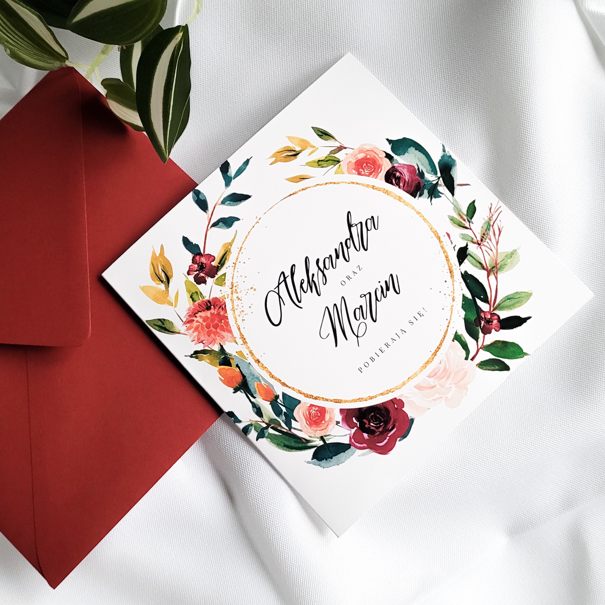 Kwadratowe zaproszenie ślubne z kwiatowym motywem i złotym okręgiem, obok bordowej koperty i rośliny na białym tle.