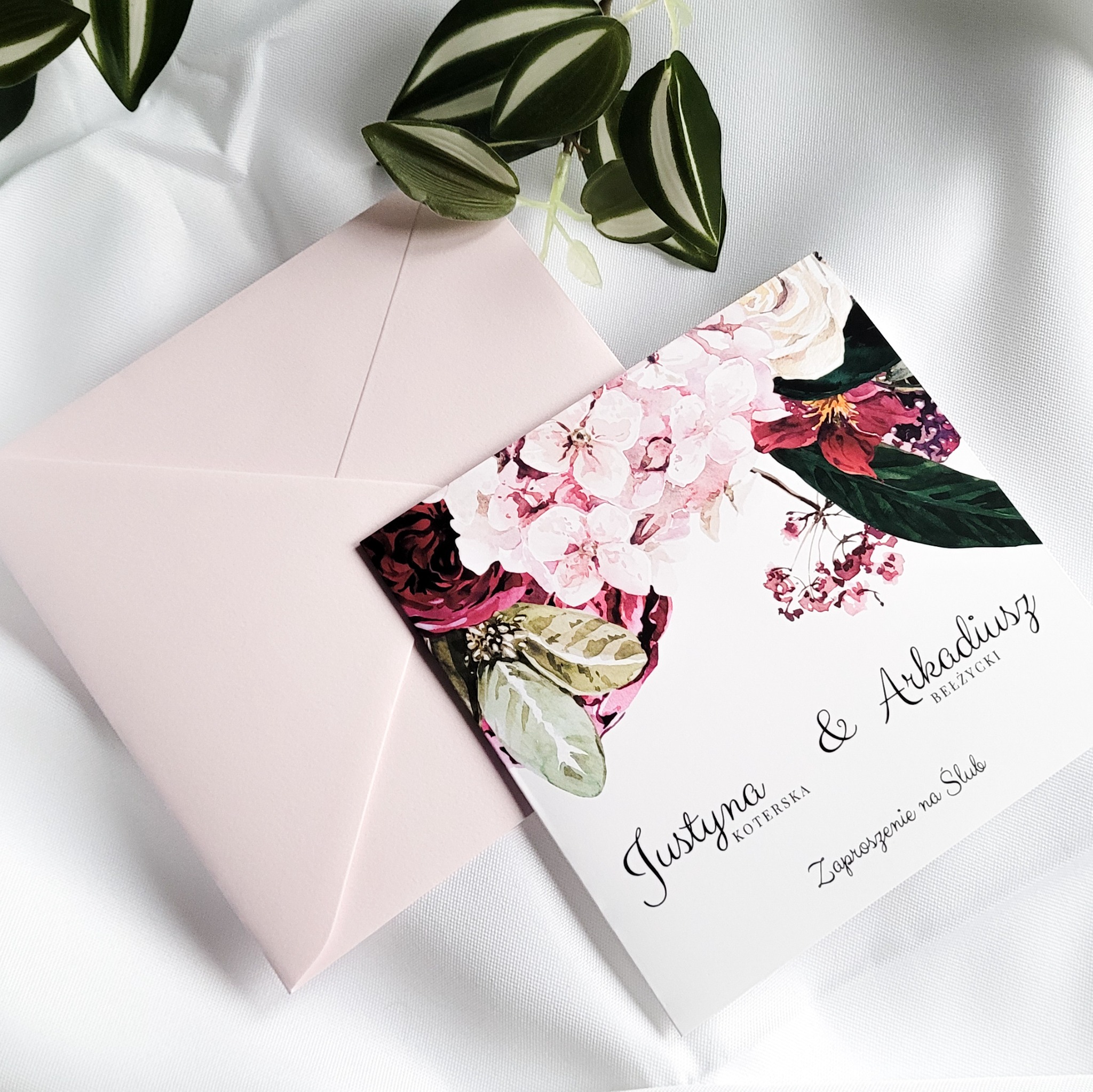Eleganckie zaproszenie ślubne z motywem kwiatowym i różową kopertą, ułożone na białej, delikatnie pogniecionej tkaninie, z dodatkiem zielonej rośliny.