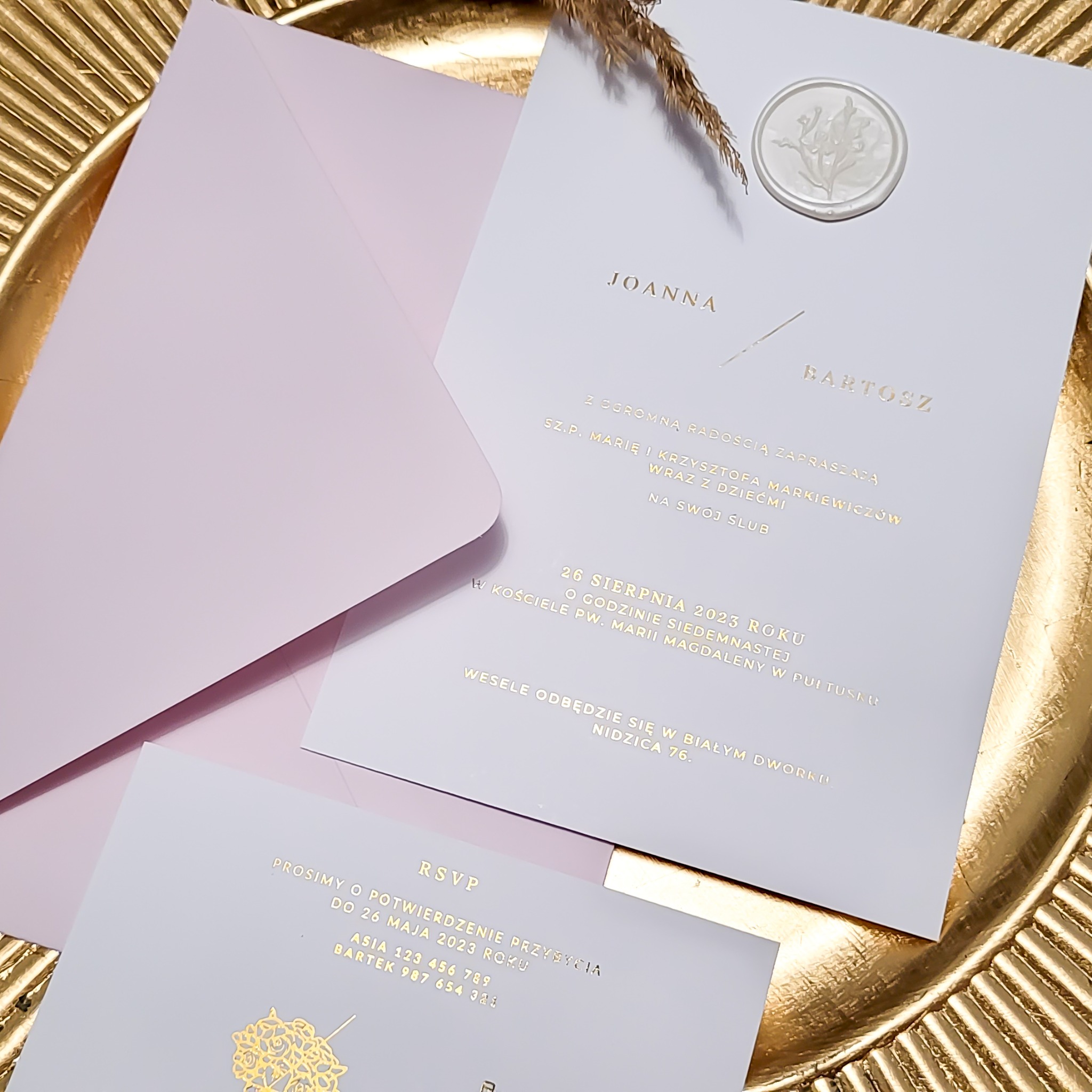 Eleganckie zaproszenia ślubne w odcieniach bieli i pudrowego różu z tłoczonym złotym tekstem, ułożone na złotej tacy dekoracyjnej, z widocznym lakowanym stemplem i suszoną trawą.