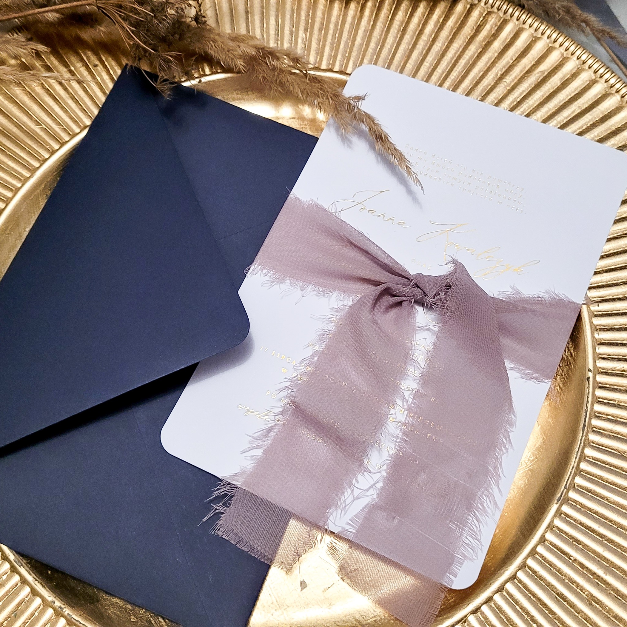 Eleganckie zaproszenie ślubne z imieniem Joanna i wstążką w odcieniach różu, leżące obok granatowej koperty na złotej paterze, z dekoracją z suszonych traw w tle.