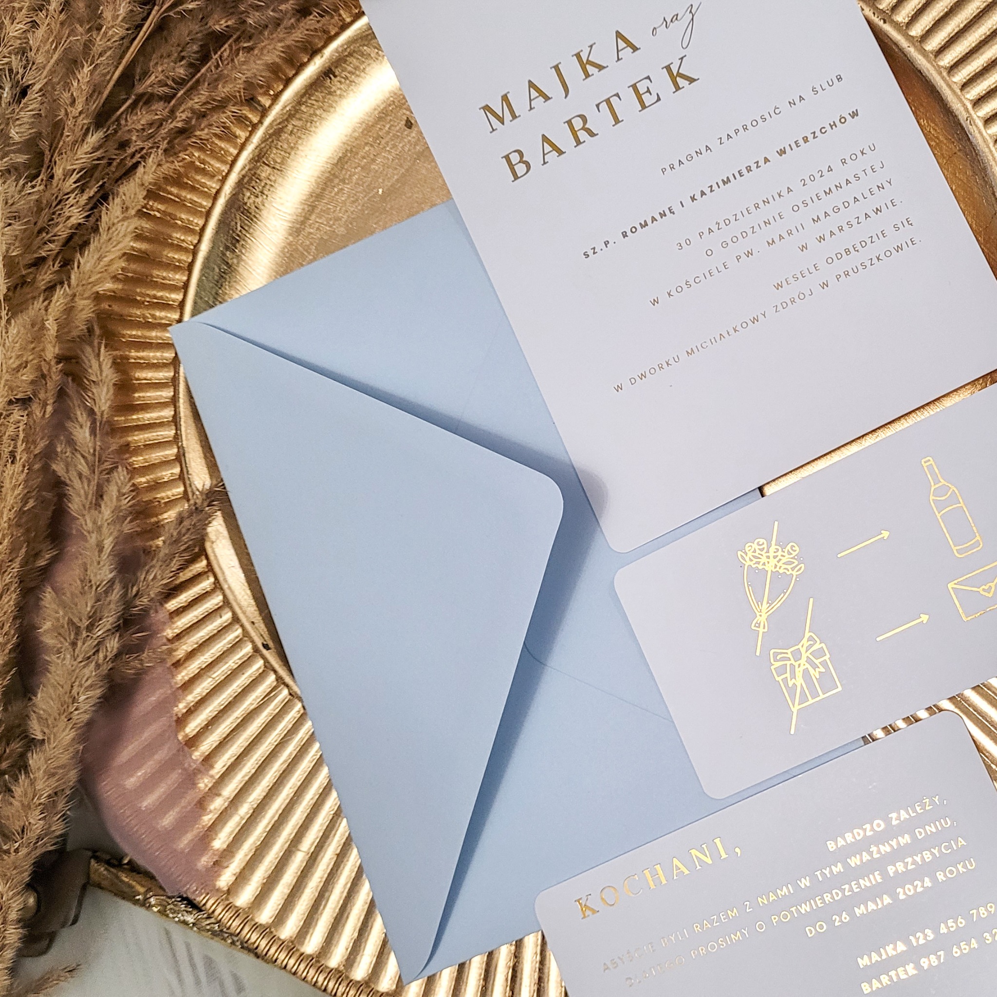 Eleganckie zaproszenie ślubne z imionami Majki i Bartka, z jasnoniebieską kopertą, leżące na złotej tacy w otoczeniu suszonych traw.