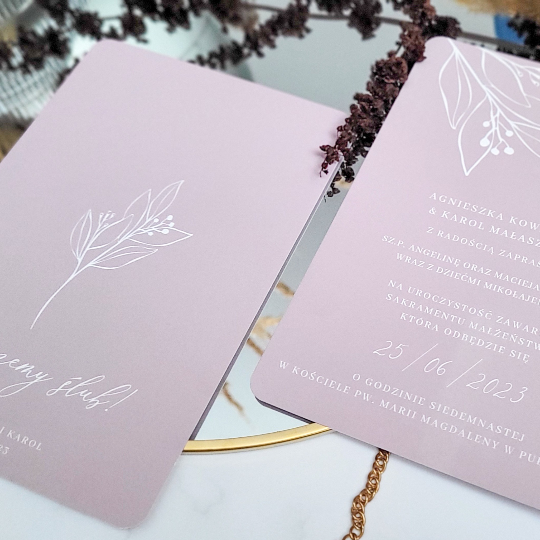 Dwa zaproszenia ślubne w odcieniach pudrowego różu z białym, minimalistycznym motywem roślinnym i tekstem, leżące na lustrzanej powierzchni z elementami złotej biżuterii i suszonych kwiatów w tle.