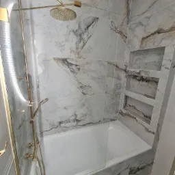 Wykończona łazienka z wanną obłożoną płytkami imitującymi marmur, z zabudowaną szklaną ścianką prysznicową z elementami w kolorze złotym oraz wnękami w ścianie.