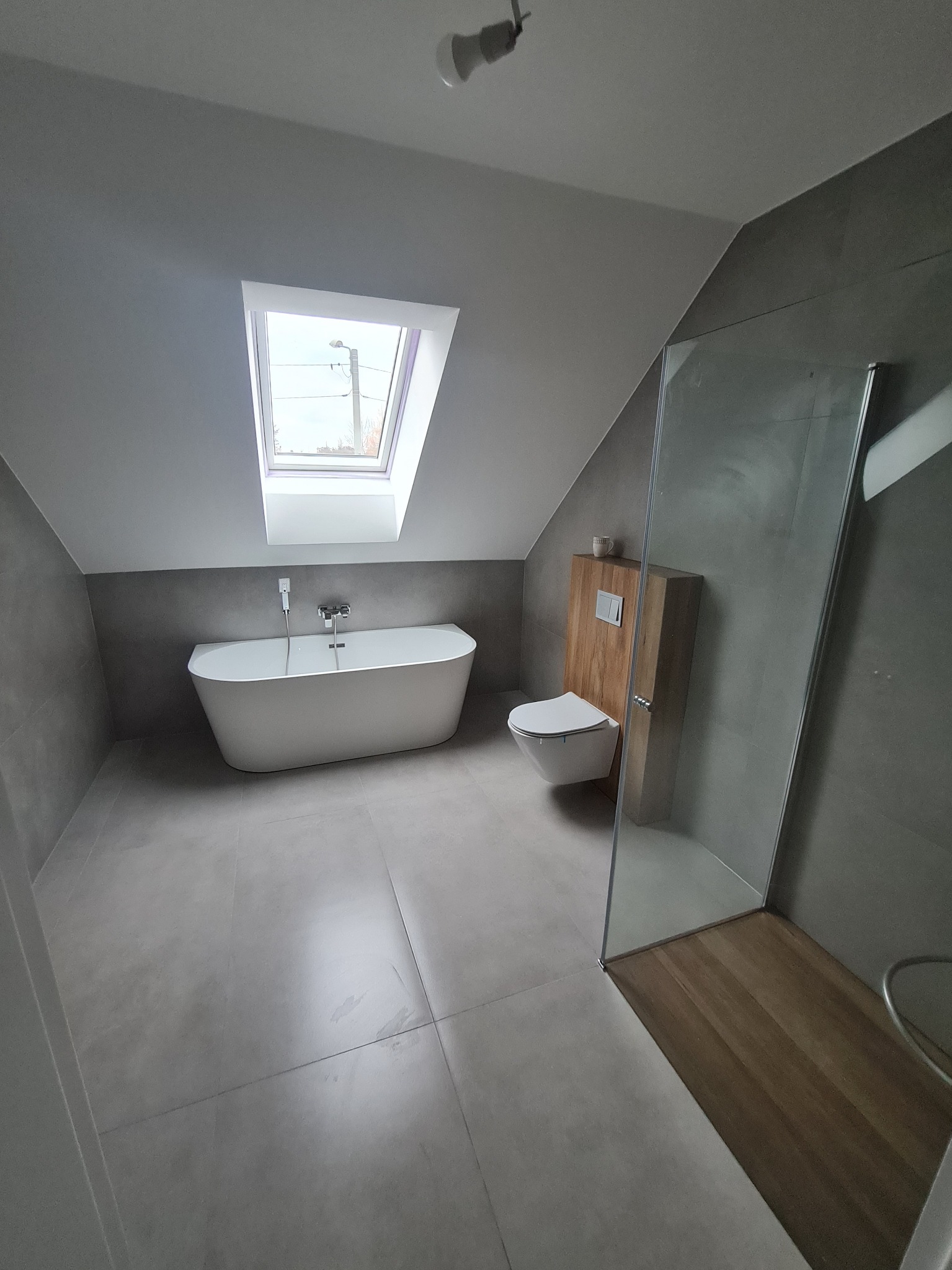 Nowoczesna łazienka na poddaszu w Olsztynie z wolnostojącą wanną, szarą glazurą ścienną i podłogową oraz kabiną prysznicową z drewnianym wykończeniem brodzika.