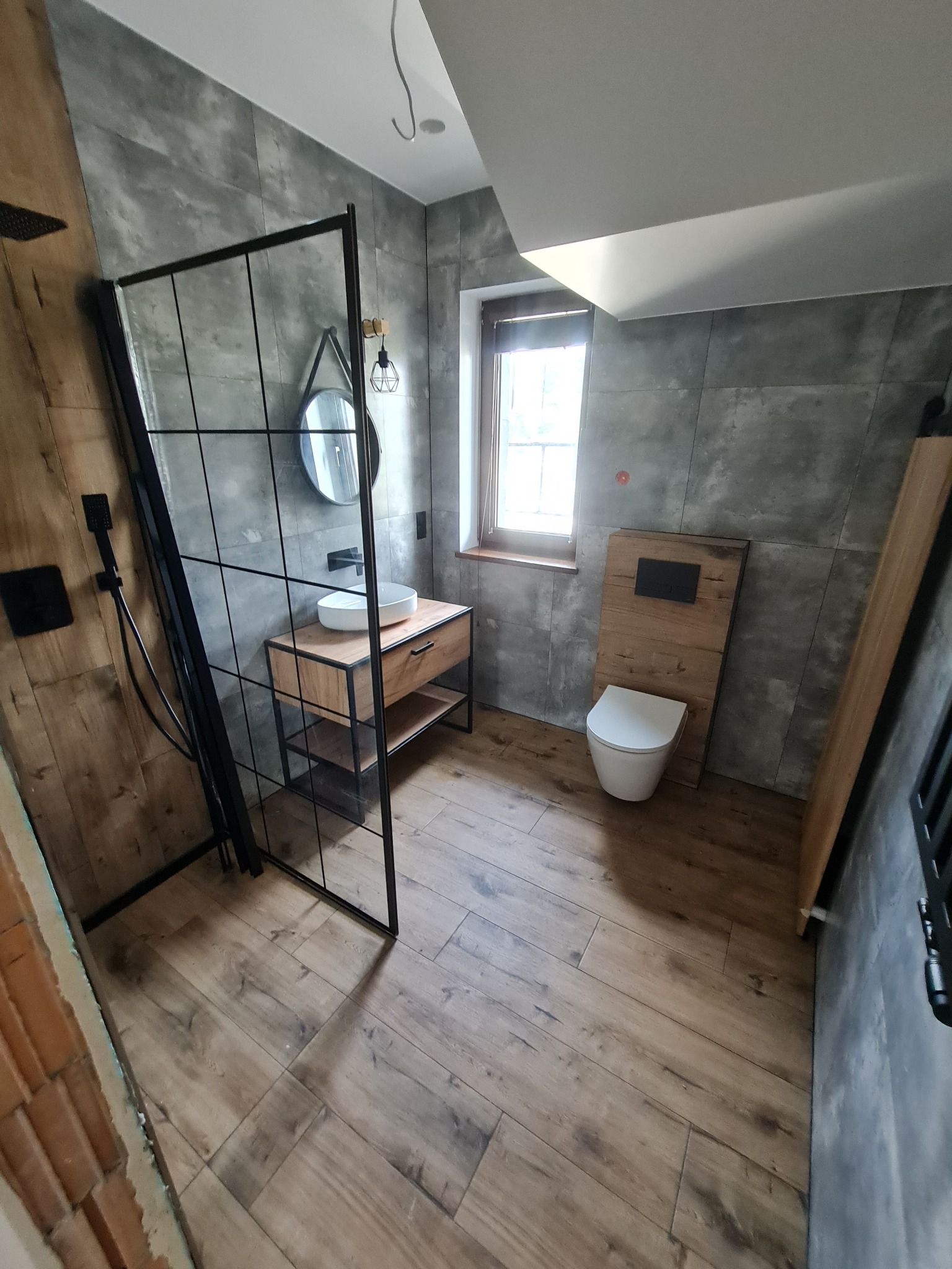 Nowoczesna łazienka z szarymi płytkami imitującymi beton, drewnianą podłogą, kabiną prysznicową typu walk-in z czarnymi profilami, okrągłym lustrem i toaletą z drewnianym panelem.